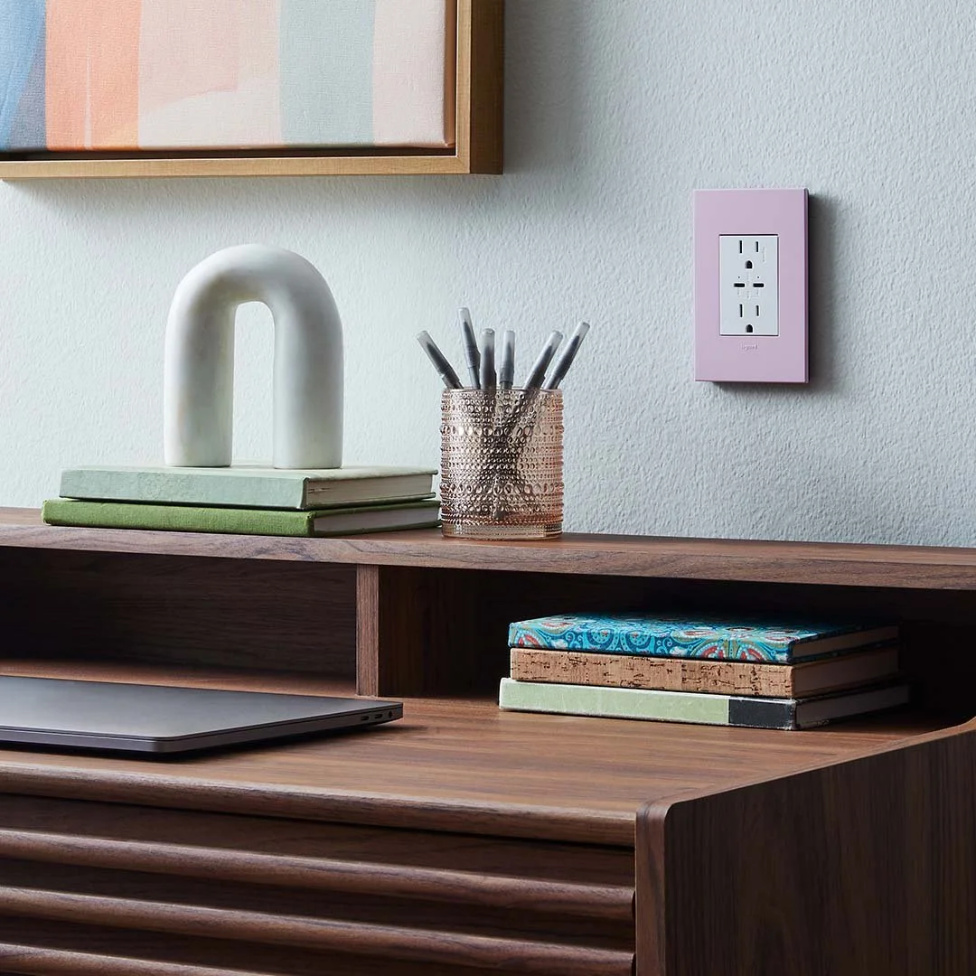 Close-up of a wooden desk with a closed laptop, a stack of notebooks, a decorative ceramic object, a metallic pen holder with pens, and a pink electrical outlet on a wall. Part of a colorful framed artwork is visible at the top of the image.