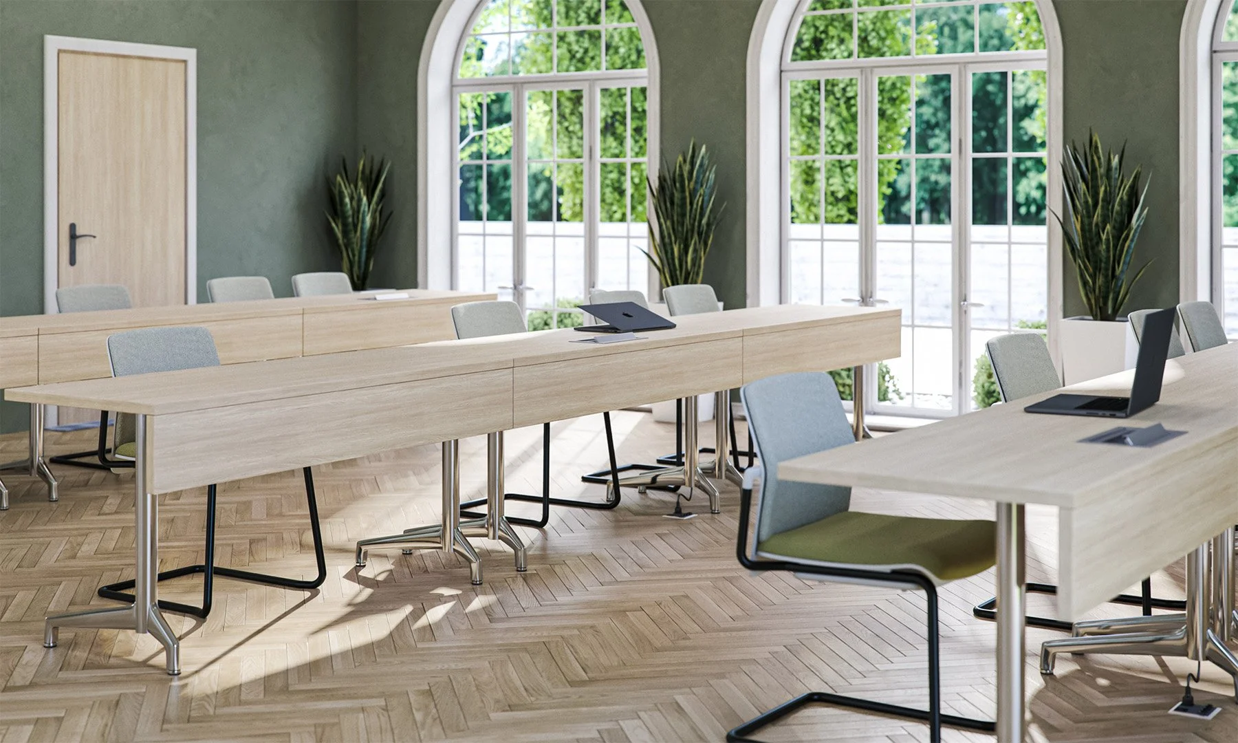 A bright conference room with large arched windows overlooking greenery, wooden flooring, and light-colored tables and chairs. There are laptops and a conference phone on the tables, and tall potted plants are placed along the windows.