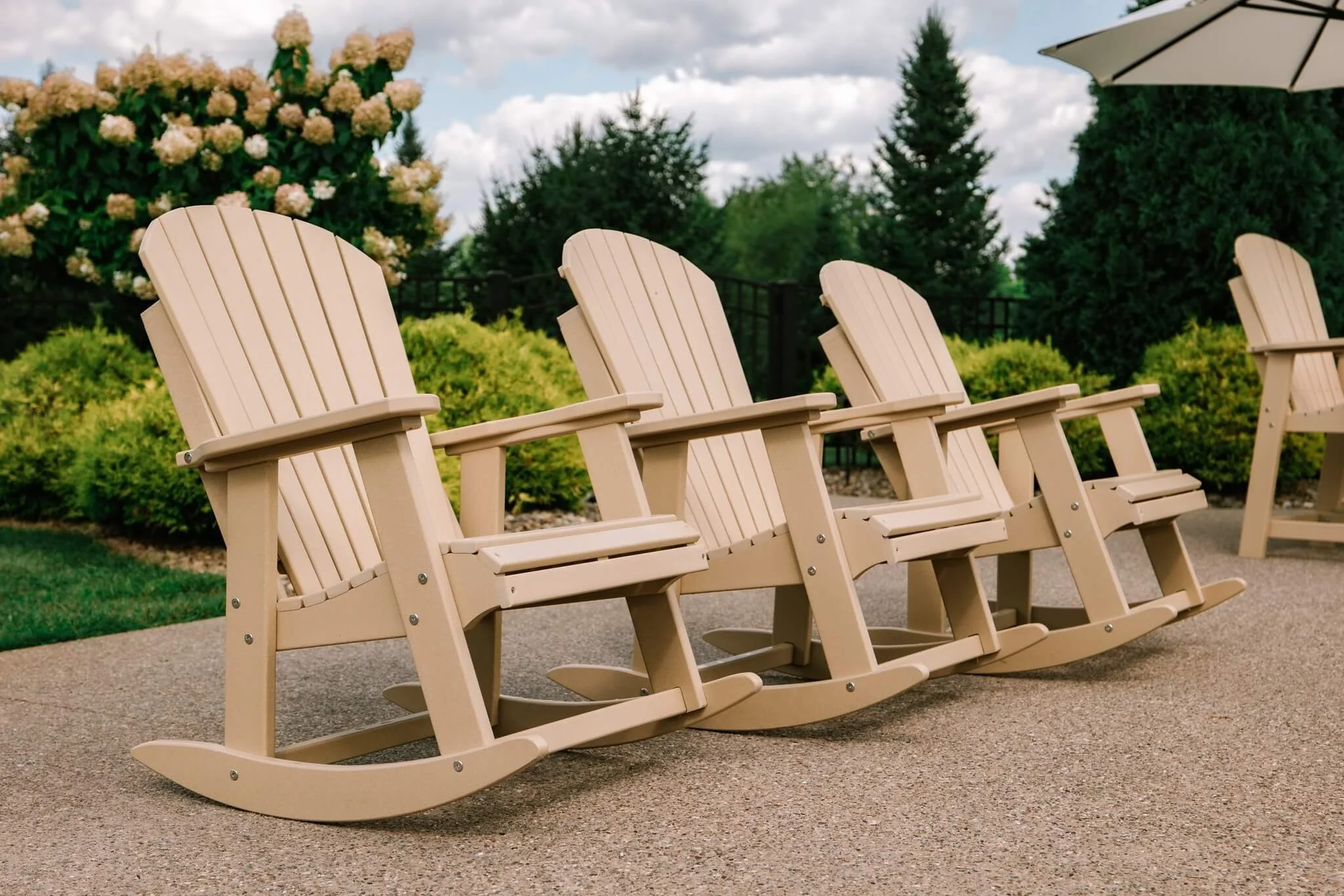 Four beige wooden rocking chairs on a concrete patio surrounded by green shrubs, trees, and flowering bushes, with umbrellas providing shade and a cloudy sky above.