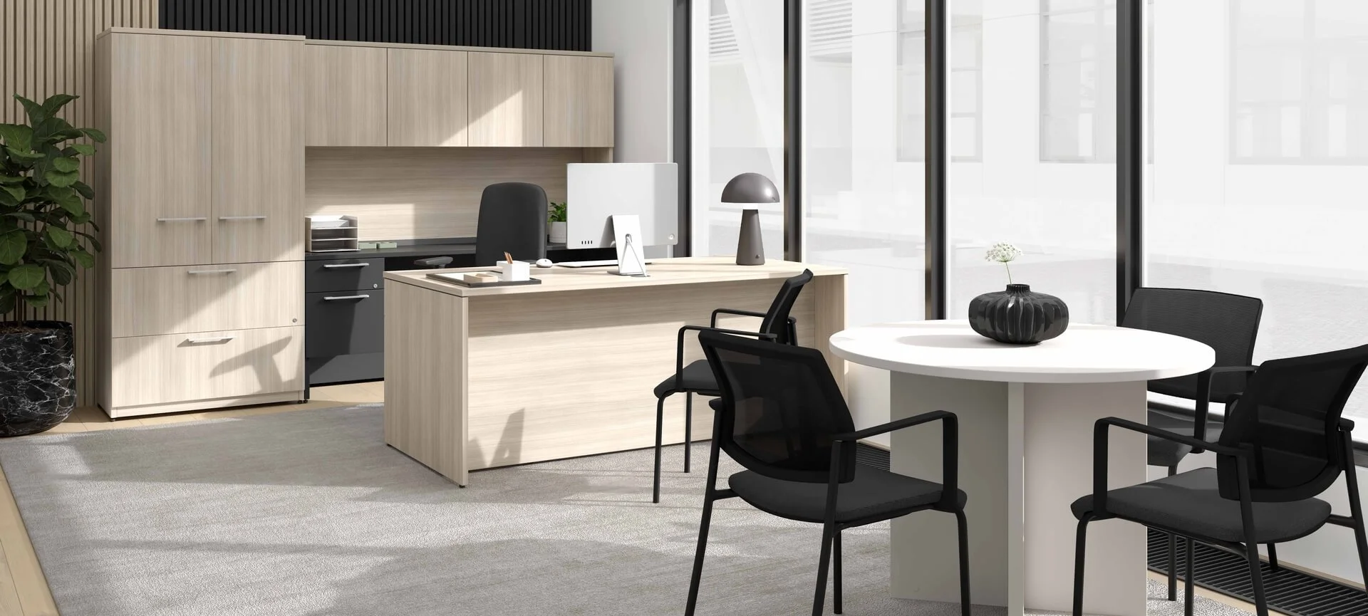Modern office space with a light wood desk, black chairs, a white round table with a black vase, large windows with natural light, and a potted plant.