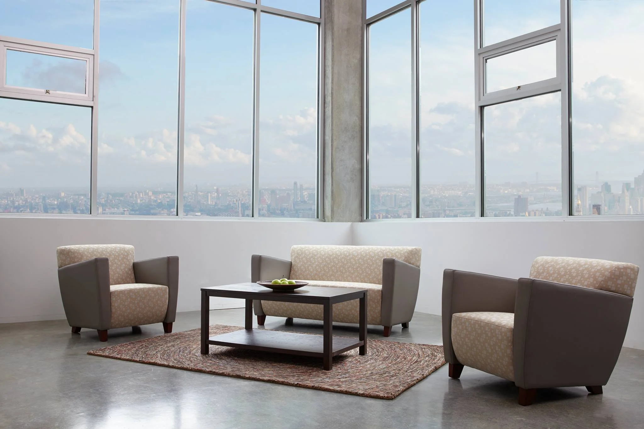 A modern living room with large floor-to-ceiling windows showing a city skyline and blue sky with clouds. There are four beige and brown upholstered armchairs around a dark wood coffee table with a bowl of green apples. A multicolored rug is under the coffee table.
