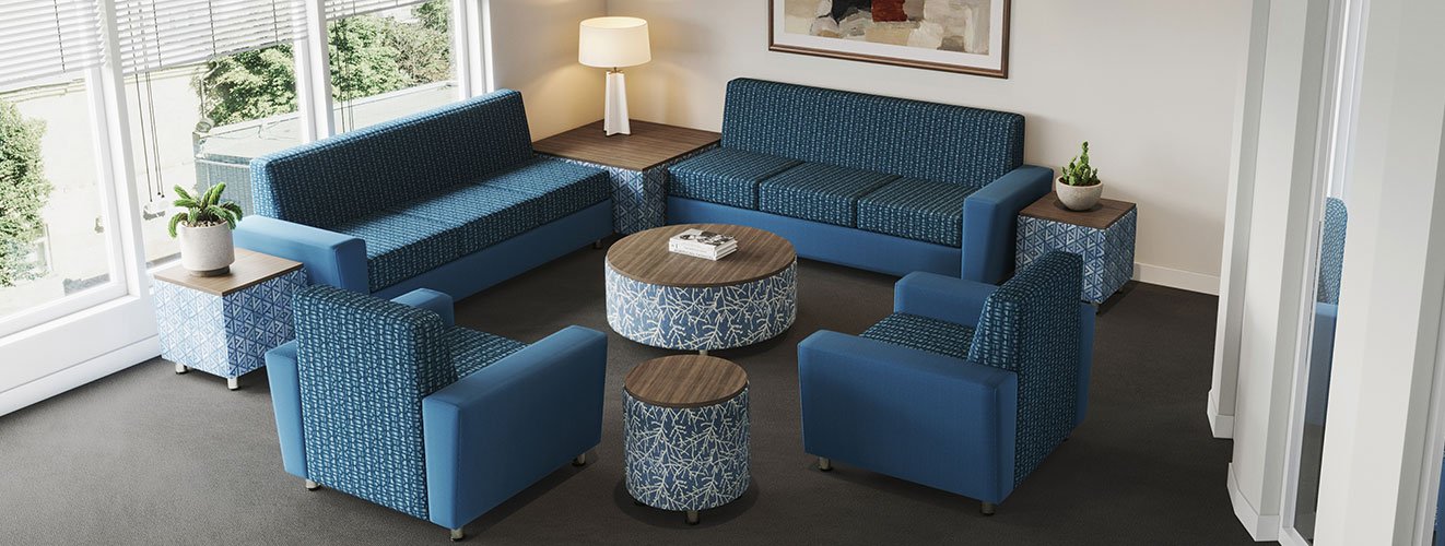 Living room with blue patterned sofas and armchairs, two side tables with potted plants, a round ottoman, and a framed painting on the wall.