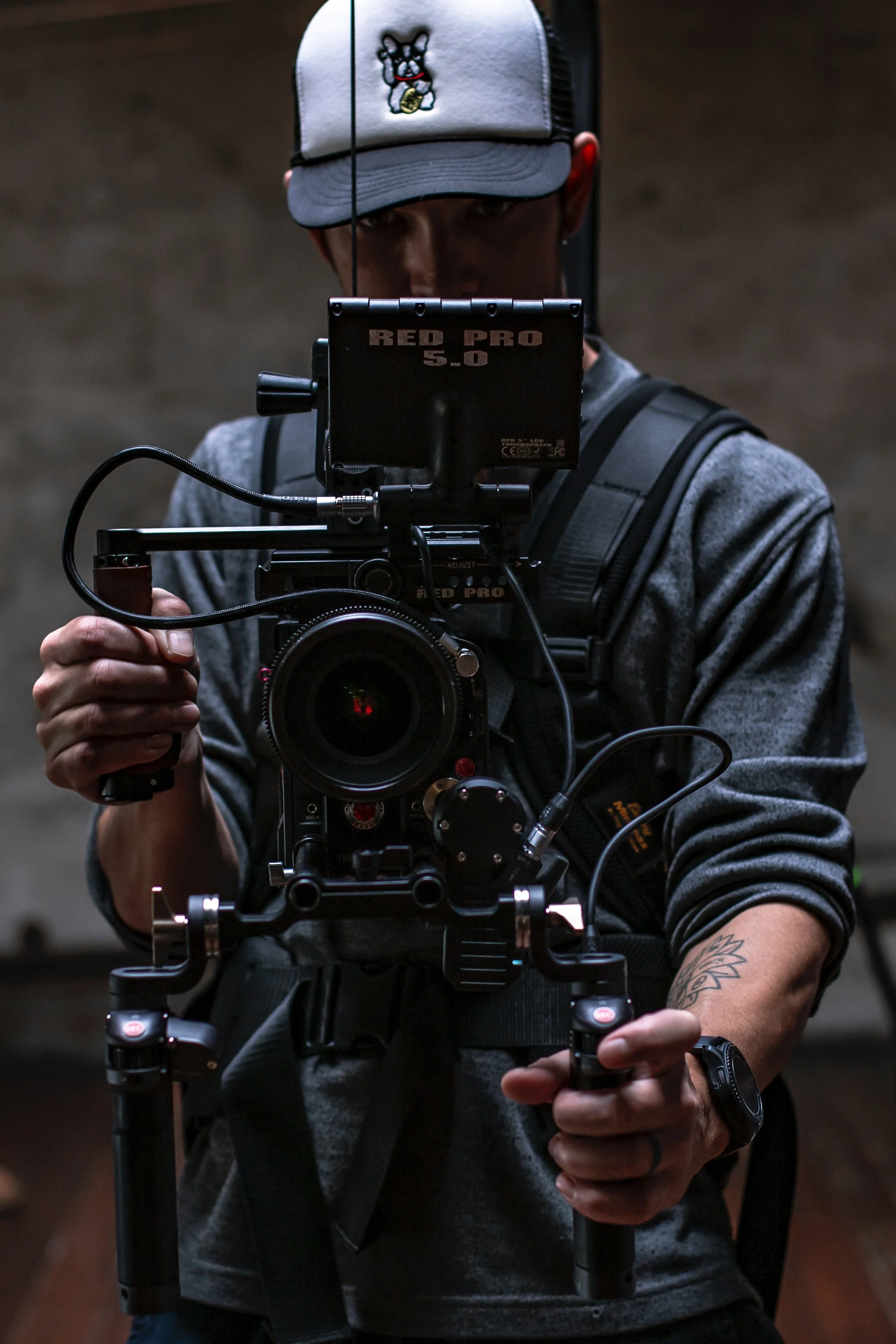 A man wearing a baseball cap and gray long-sleeve shirt operating a professional camera rig, with a blurred background.