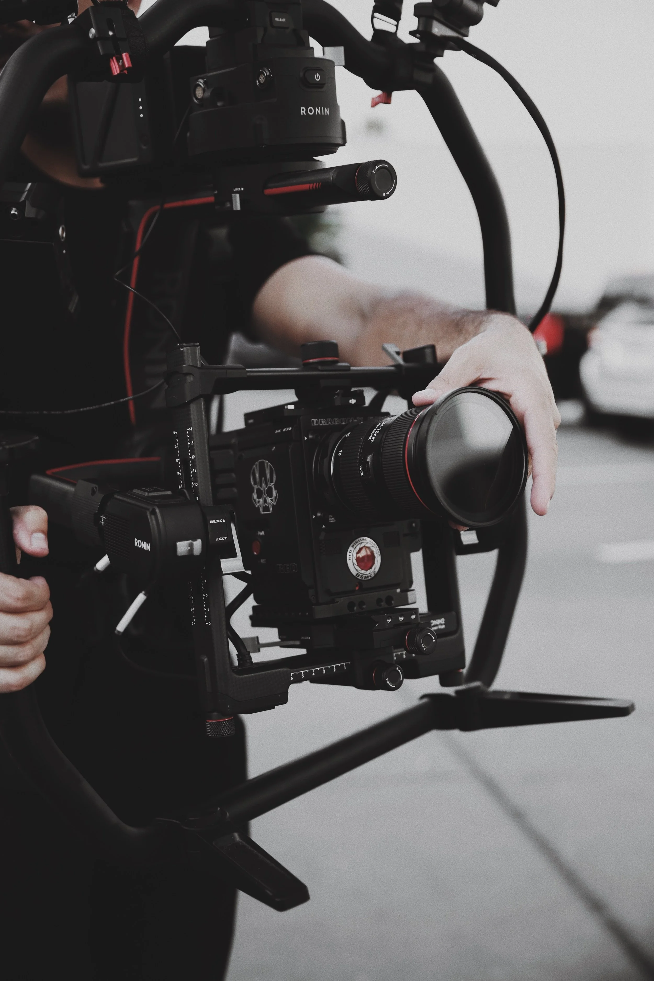 Close-up of a professional camera rig operated by a person holding the camera handle with their right hand, with various camera components and mounting hardware visible.