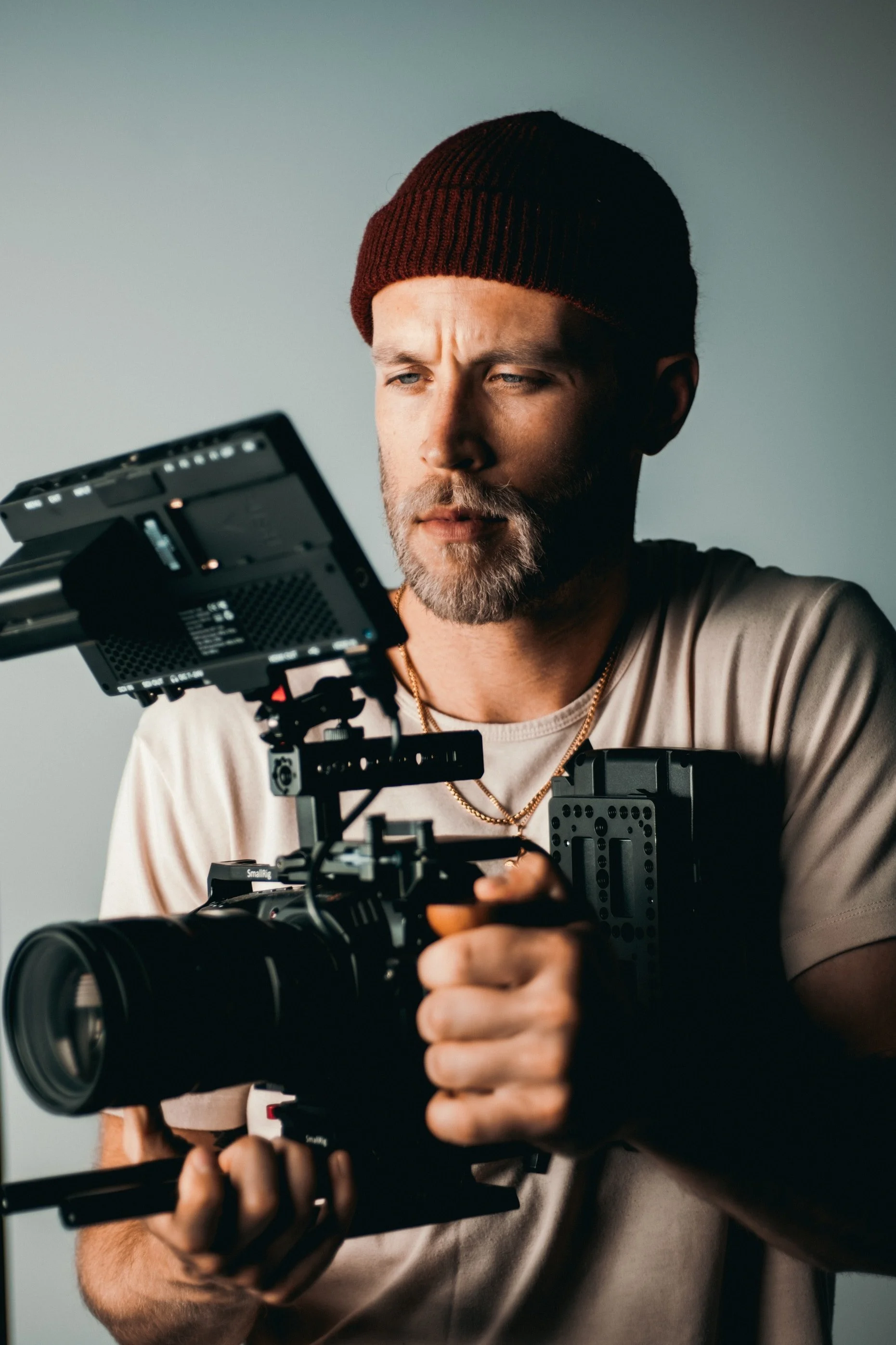 A man with a beard wearing a maroon beanie and beige t-shirt operating camera equipment.