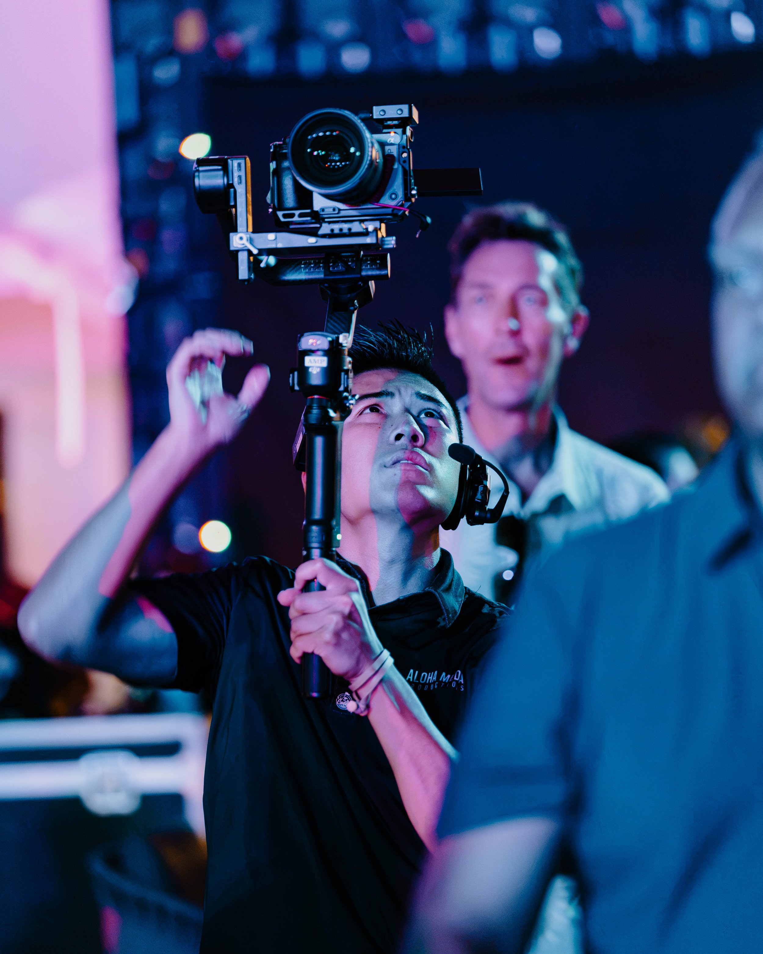 A man wearing a baseball cap and gray long-sleeve shirt operating a professional camera rig, with a blurred background.