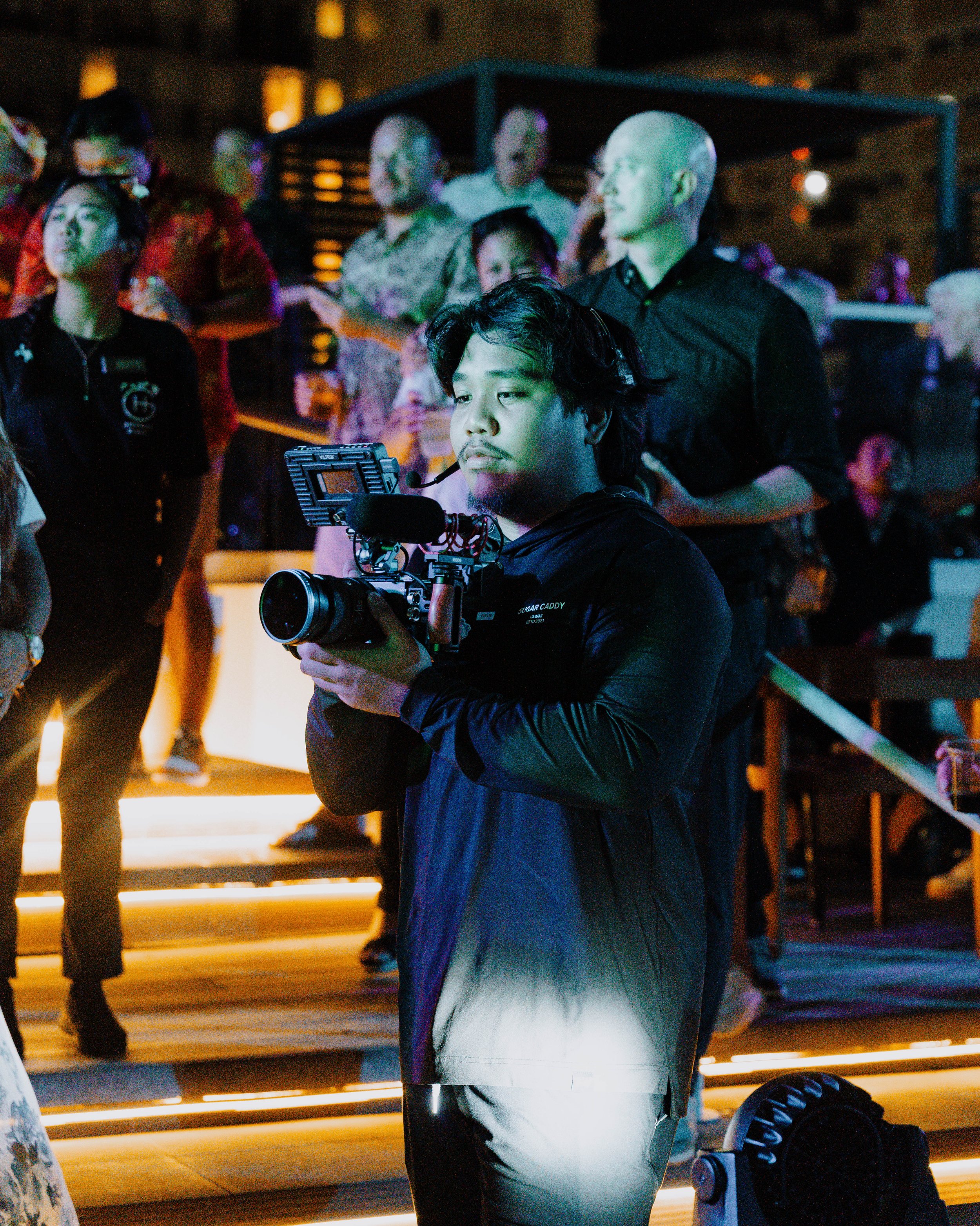 Close-up of a professional camera rig operated by a person holding the camera handle with their right hand, with various camera components and mounting hardware visible.