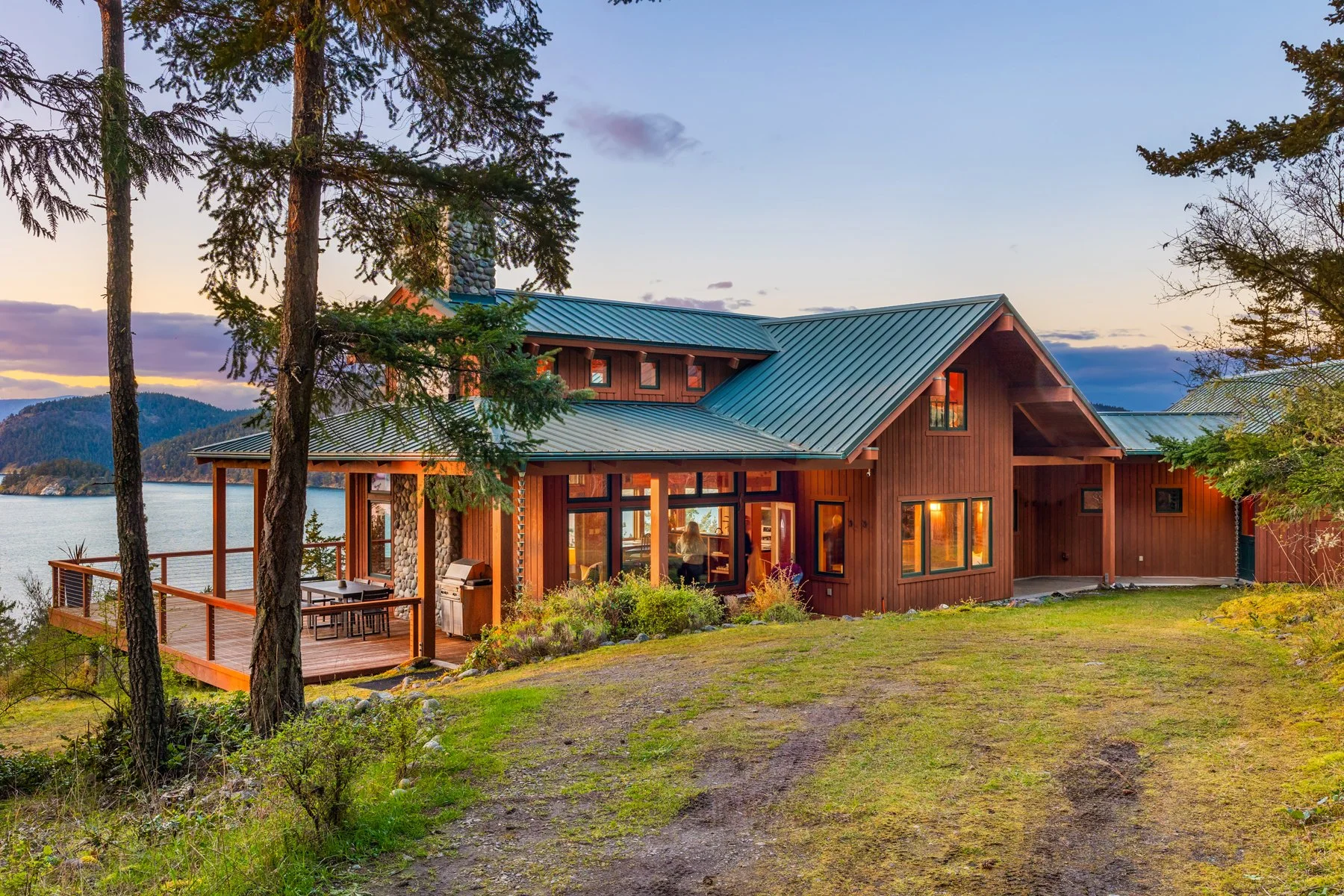 A modern lakeside home with a green metal roof, large windows, and a wooden deck overlooking a body of water, surrounded by trees and a grassy area at sunset.