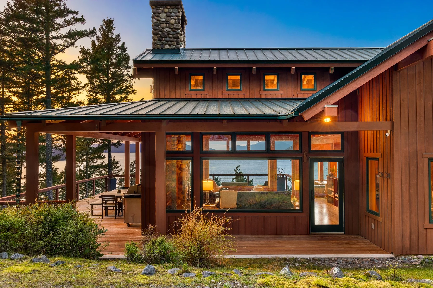 A wooden house with a metal roof, large windows, and a chimney, set in a forested area during sunset, overlooking a body of water.