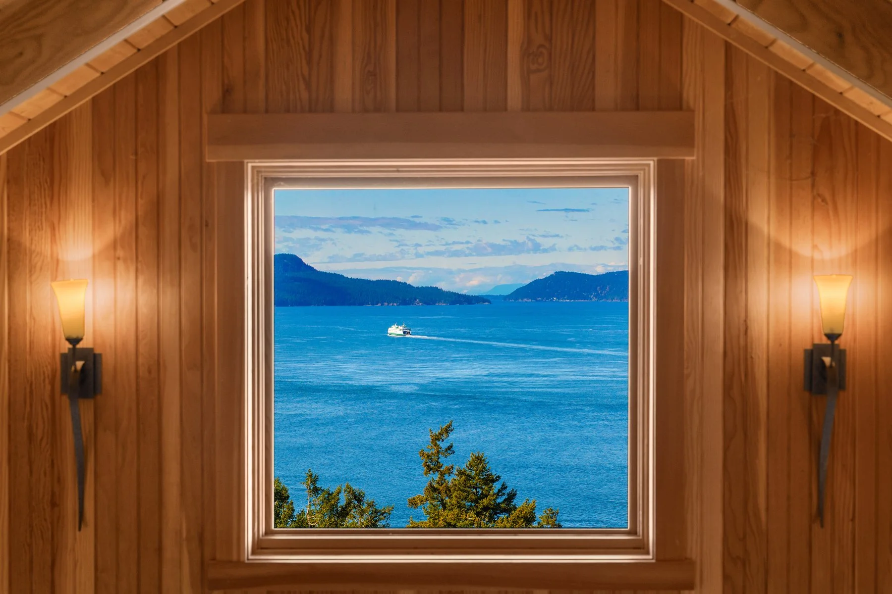 A view of a large body of water with mountains in the distance as seen through a window in a wooden room, with two wall sconces on either side.