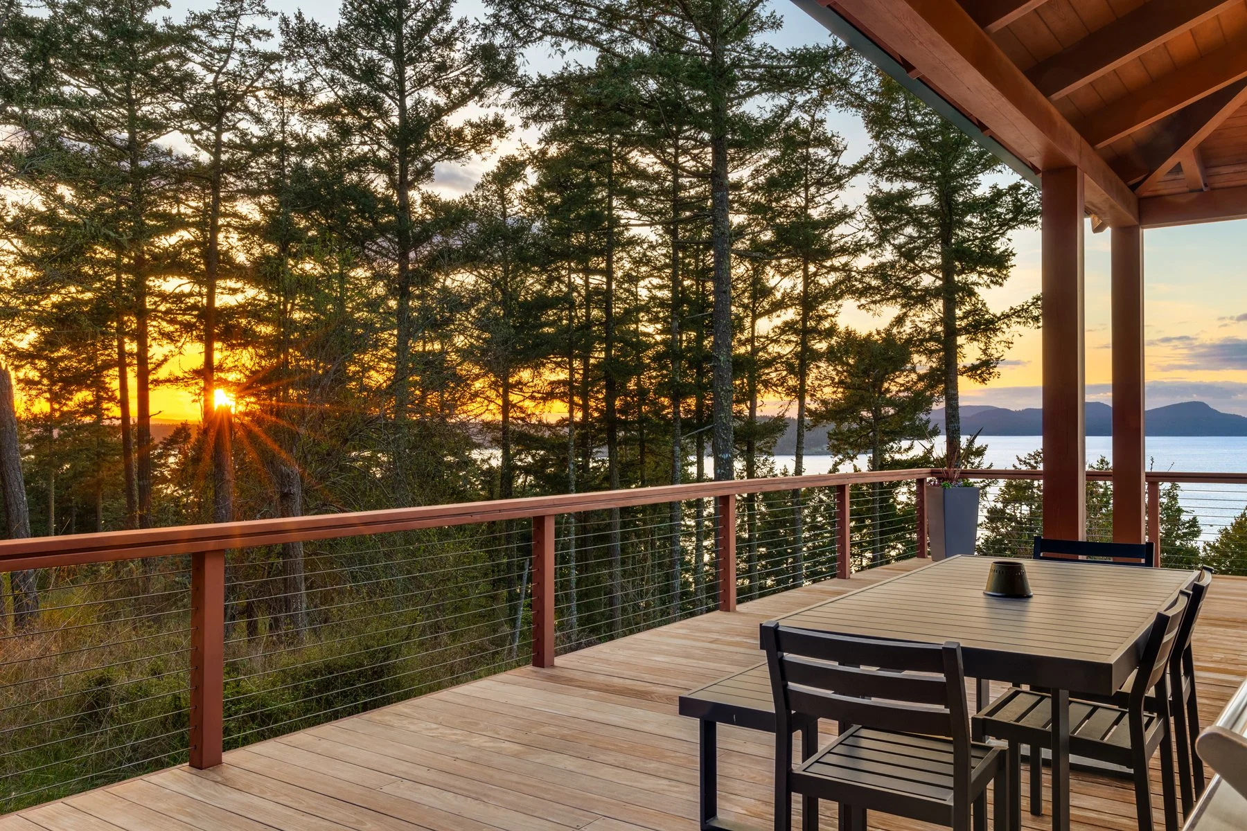 Sunset view from a wooden deck overlooking a forest of trees with mountains and a body of water in the distance.
