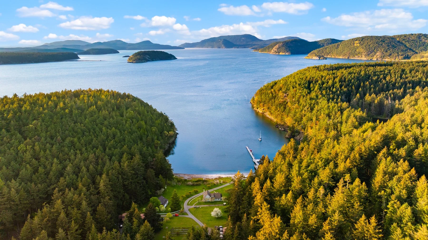 A scenic view of a lake surrounded by green and yellow forested hills under a partly cloudy sky, with a small dock and boats near a house at the bottom.