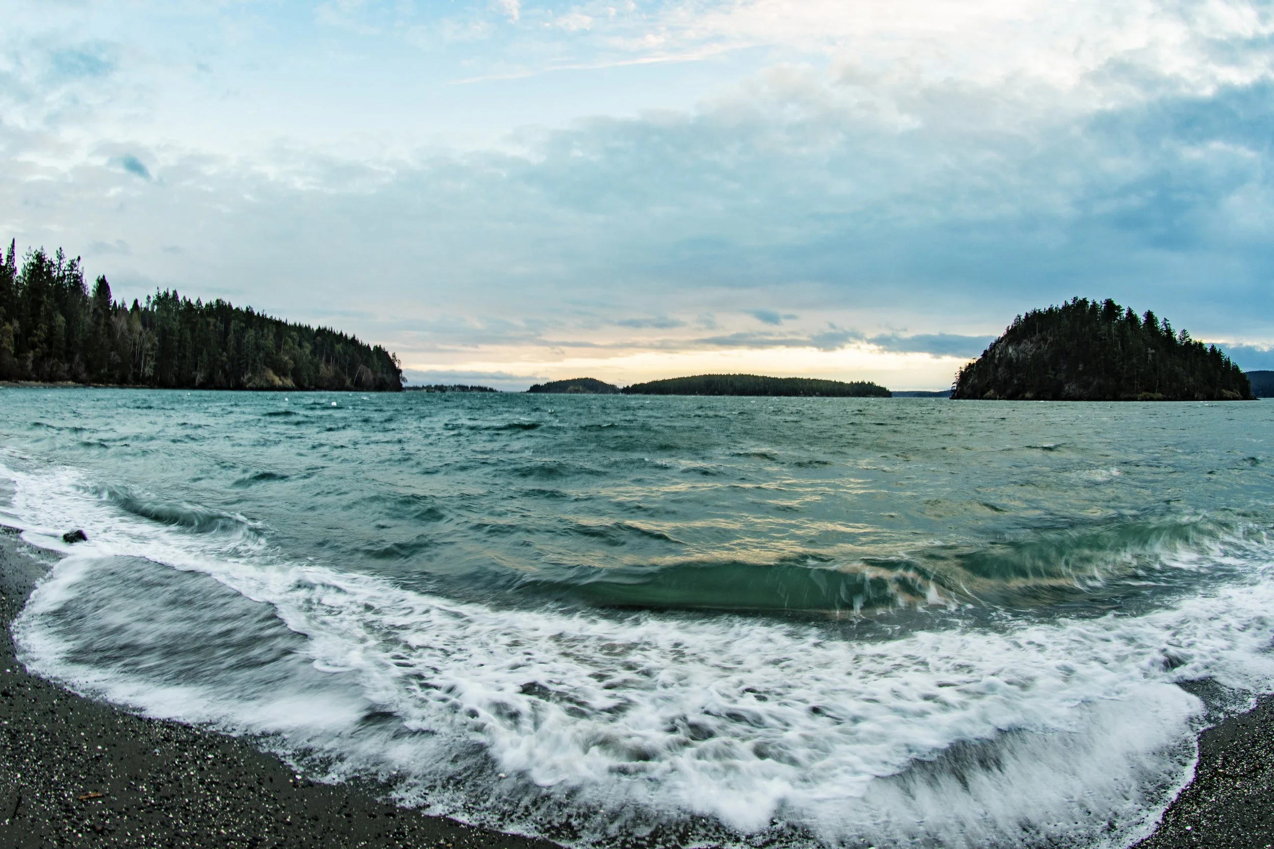 A body of water with a rocky shoreline in the foreground, surrounded by forested islands and a cloudy sky.