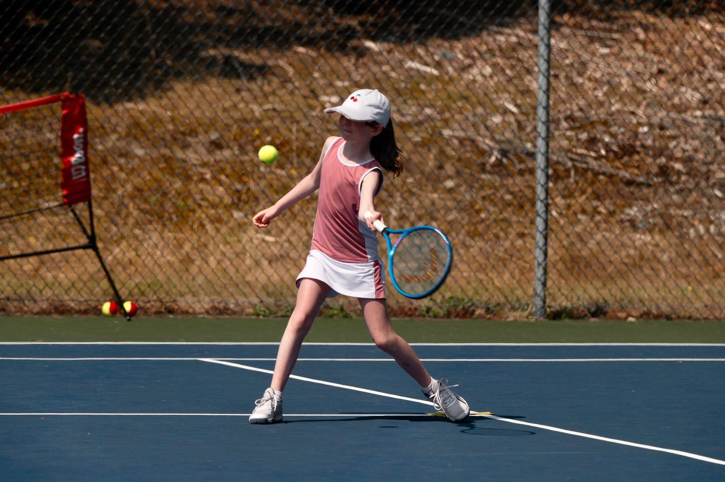 Young girl playing tennis on a court, wearing a white cap, pink sleeveless top, and white skirt, about to hit a tennis ball with a blue racket.