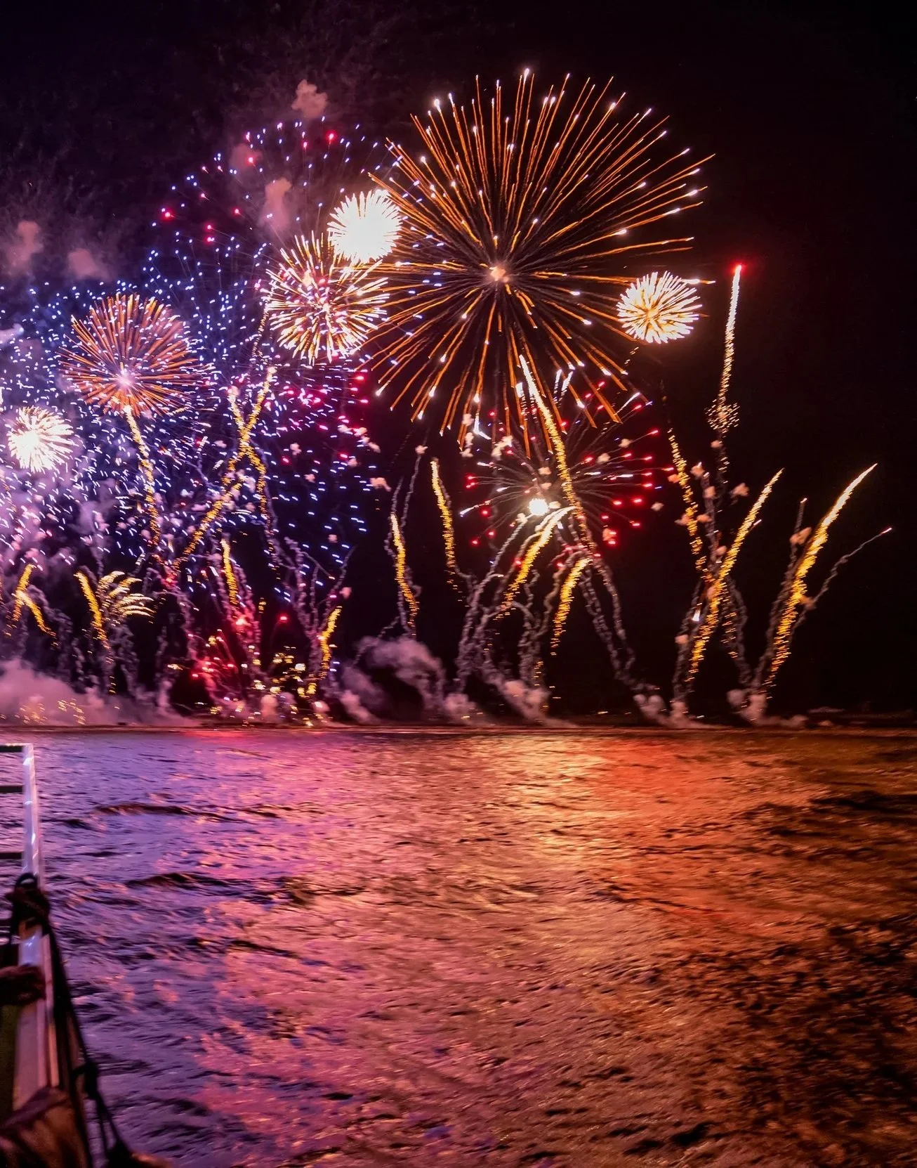 Colorful fireworks lighting up the night sky over a body of water, with reflections of the fireworks visible on the water's surface.