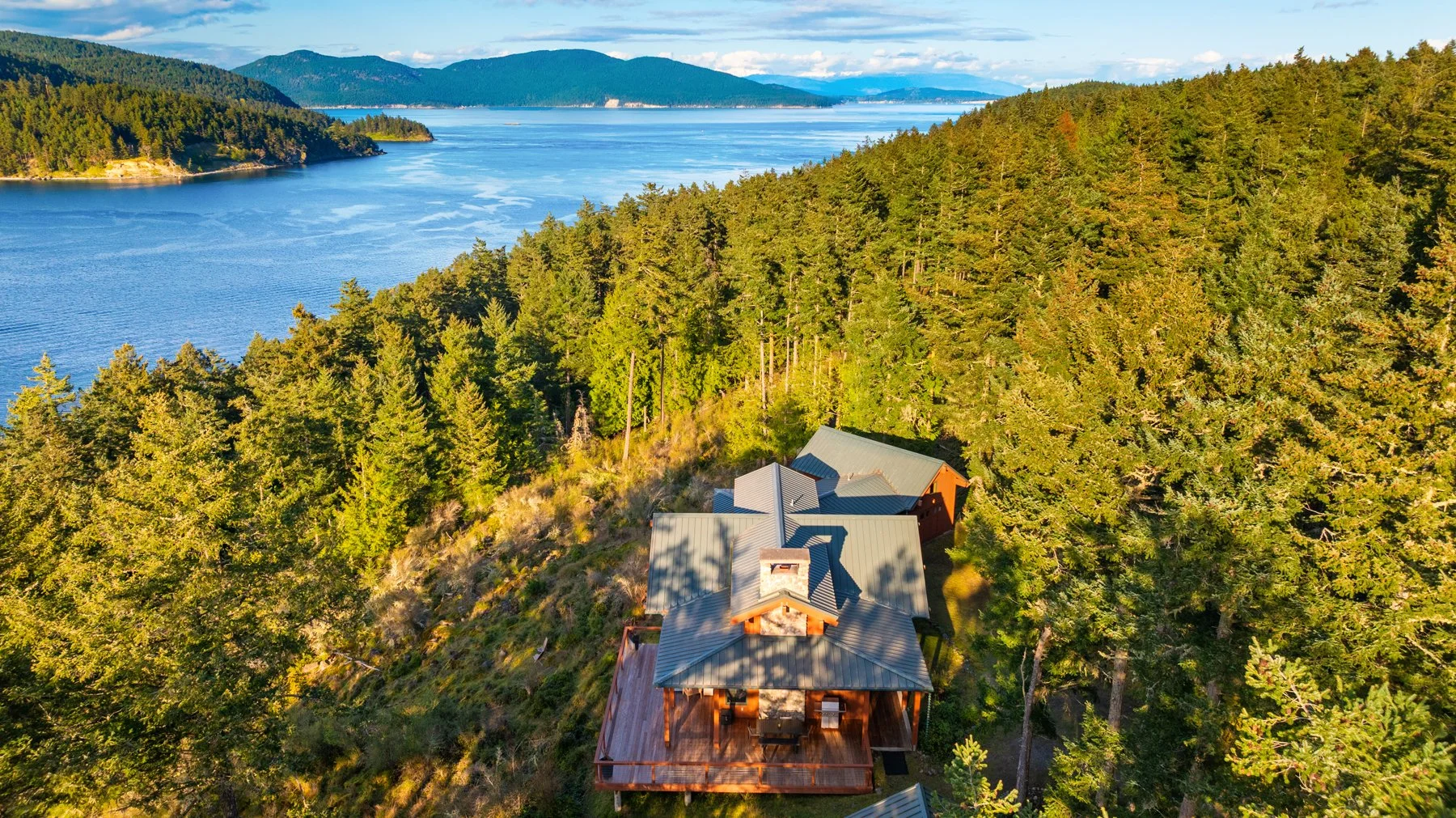 A house on a hillside surrounded by dense forest overlooking a large lake with mountains in the distance.