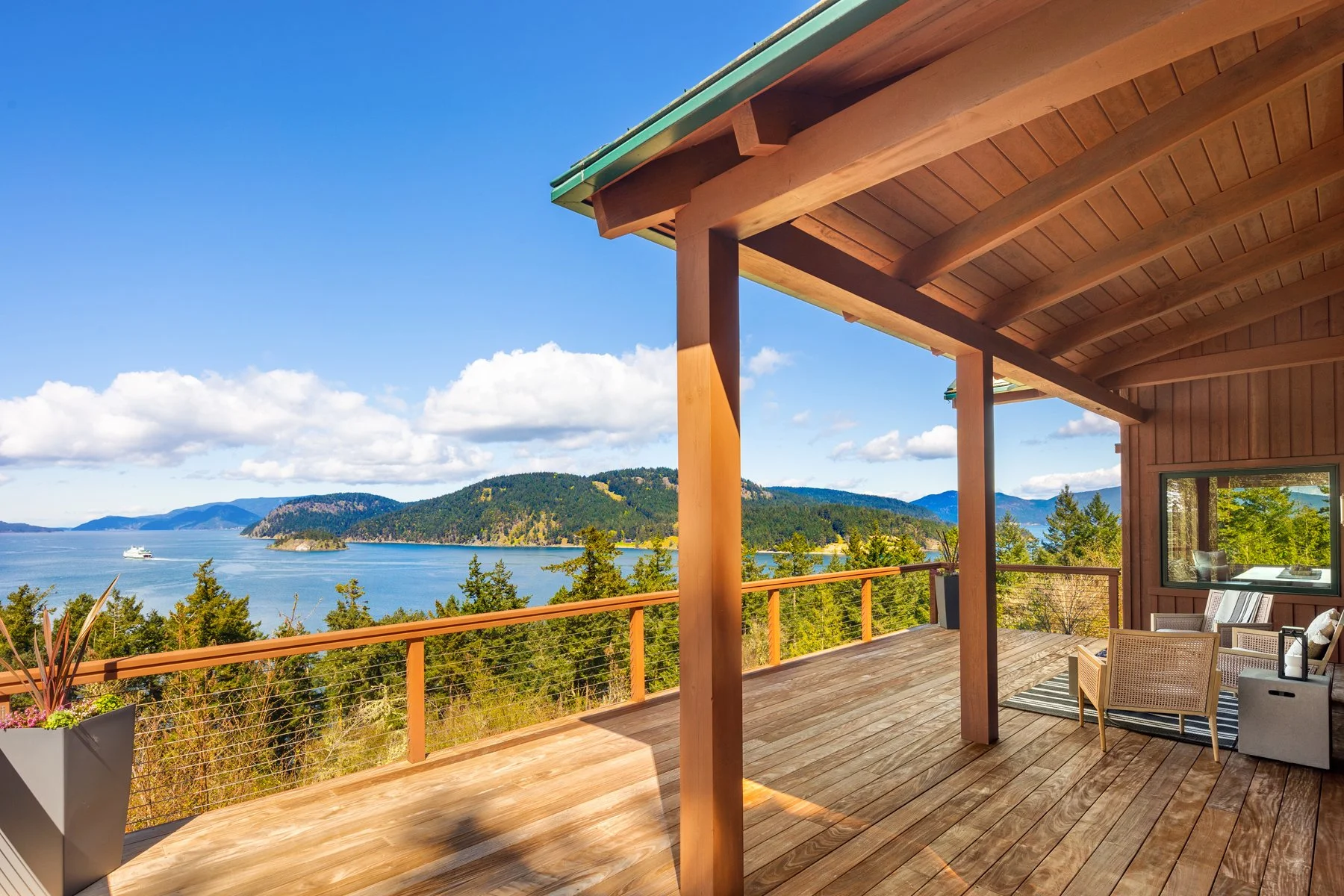 A wooden deck with outdoor seating and a view of a lake and forested mountains under a blue sky with clouds.