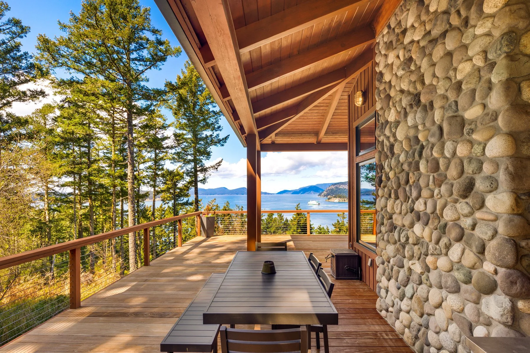 A spacious wooden balcony with a stone wall on one side, overlooking tall pine trees and a large body of water with distant mountains, under a clear blue sky.