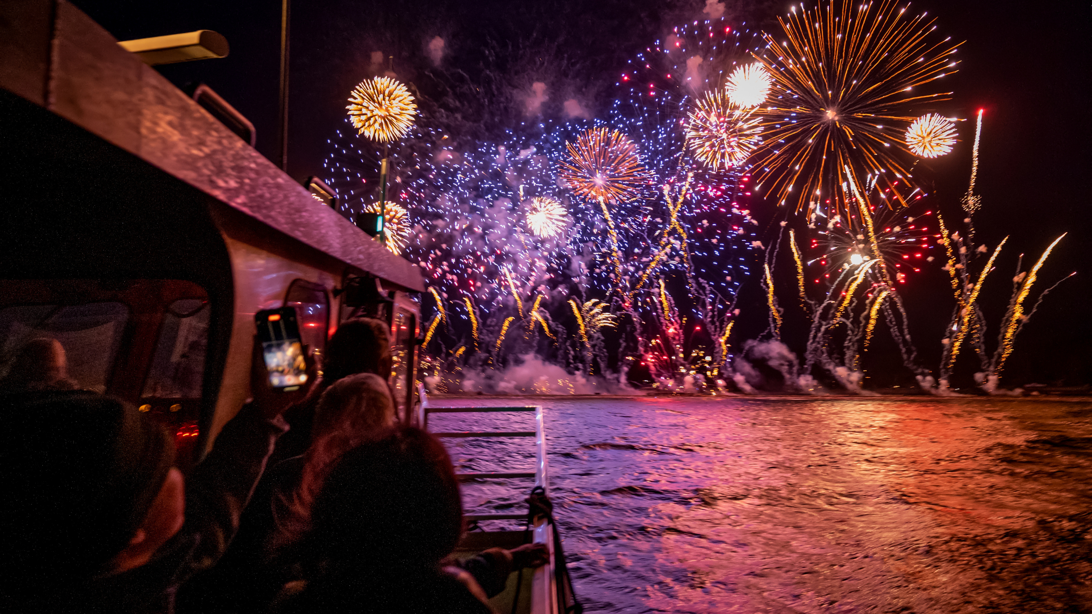 Fireworks display over water at night, with people on a boat and onshore capturing the scene.