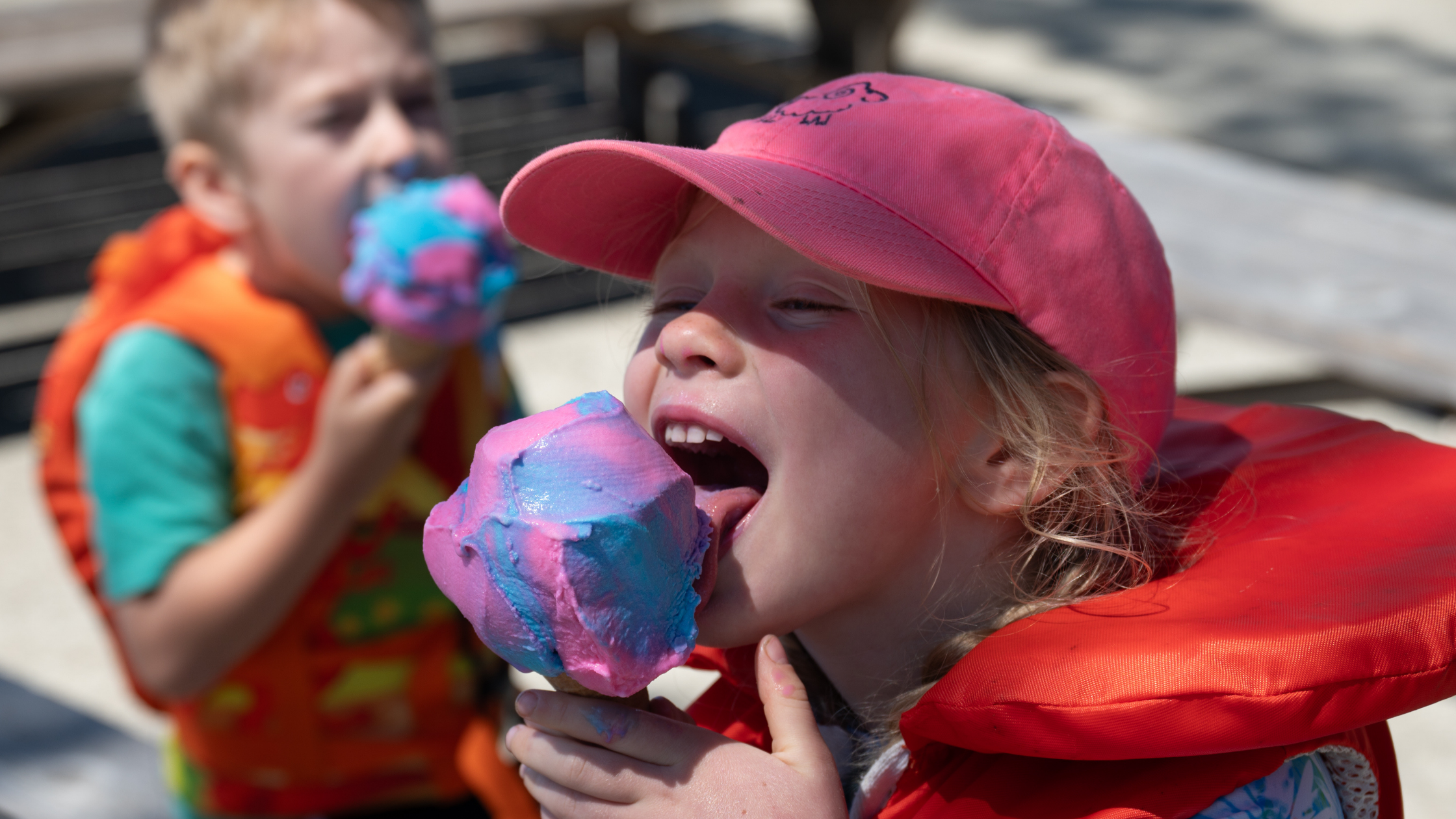 A young girl wearing a pink cap and orange life jacket eating rainbow-colored cotton candy outdoors on a sunny day, with another child in the background also holding cotton candy.