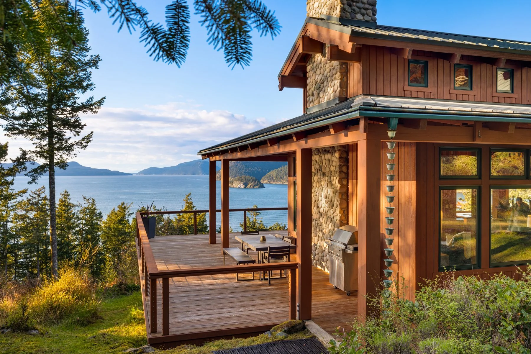 A wooden house with large windows and a stone chimney, situated on a hillside, overlooking a body of water with mountains in the distance. There is a spacious deck with outdoor furniture, surrounded by trees and greenery, under a clear blue sky.