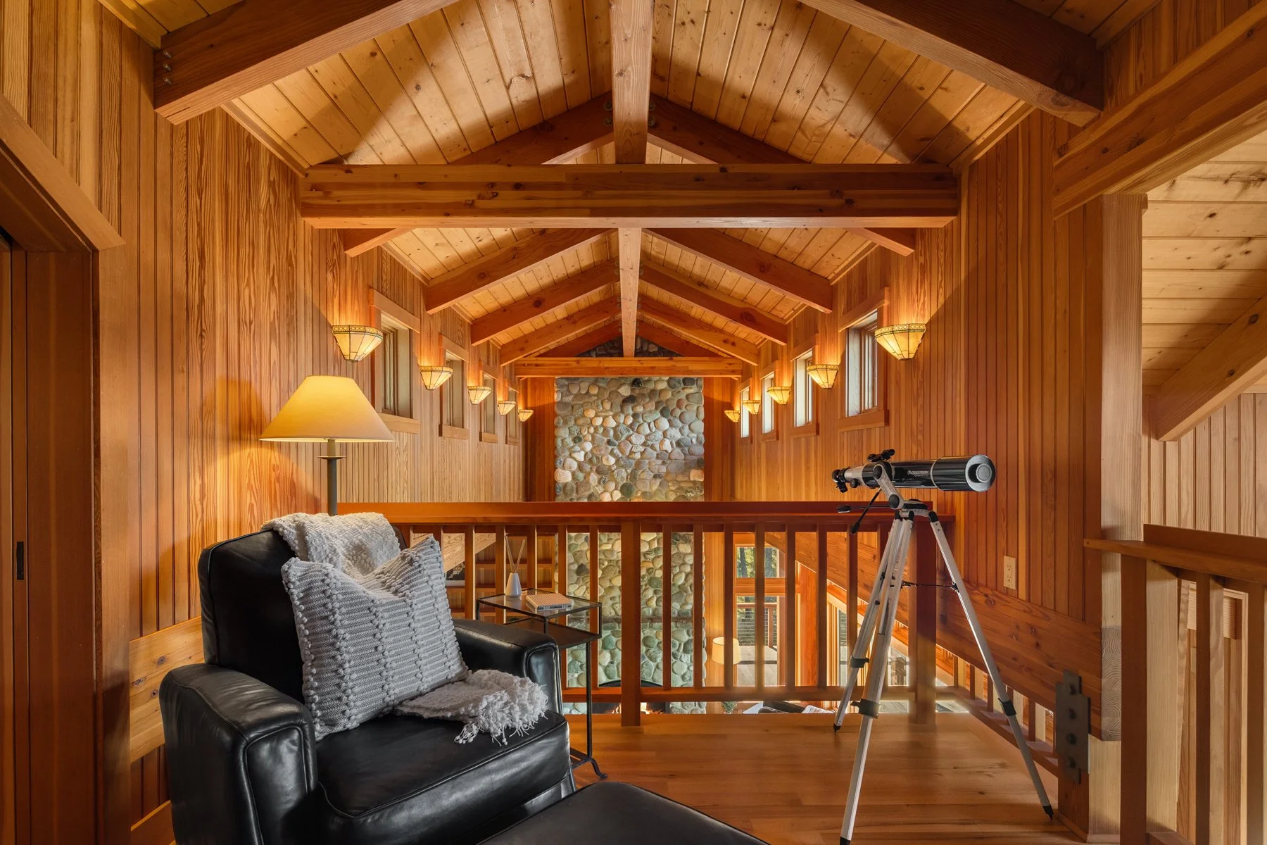 Wood-paneled upper-level room with sloped ceiling, multiple narrow windows, a black leather armchair with pillows and a blanket, a small side table with a lamp, a telescope on a tripod, and a stone wall feature in the background.