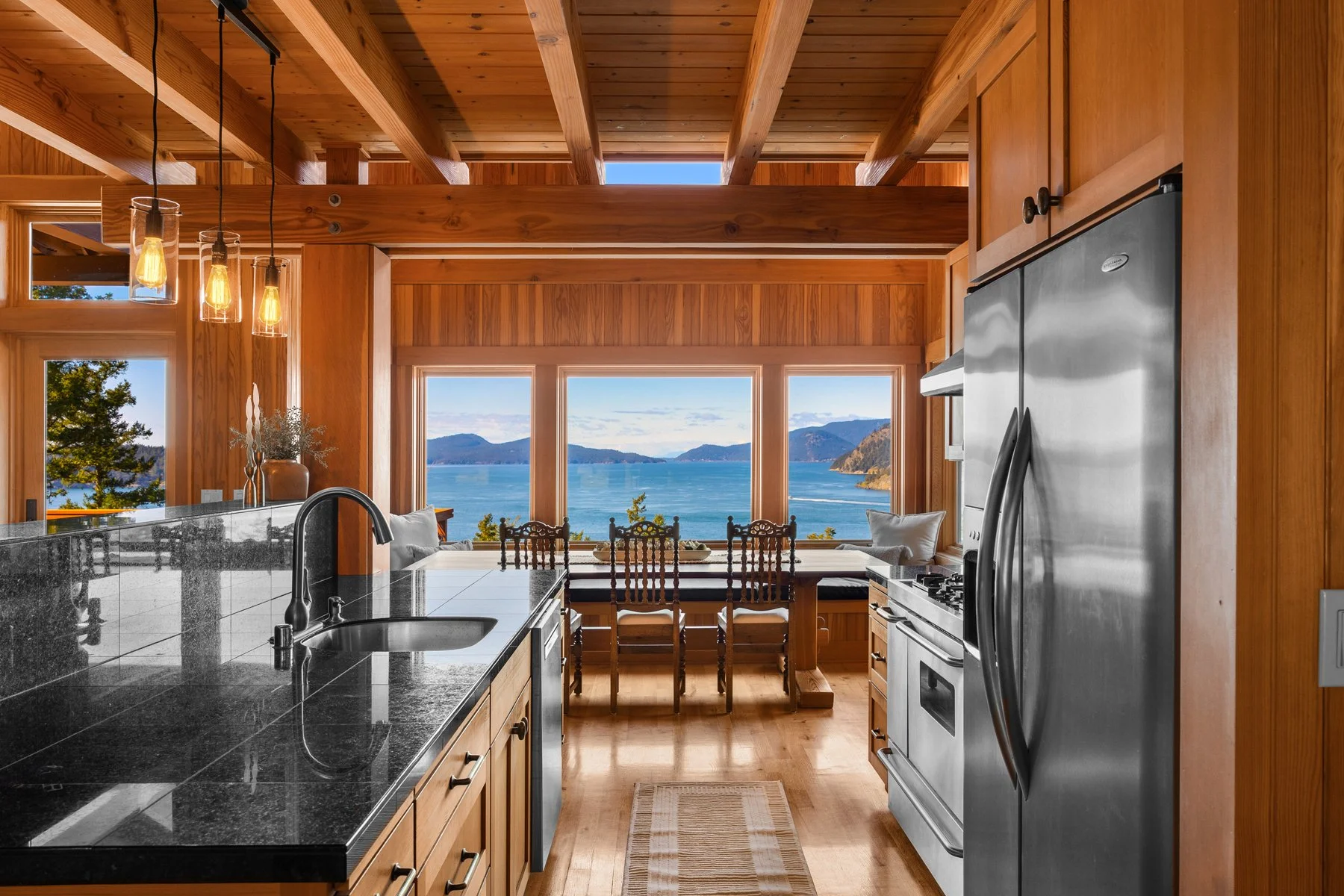 Kitchen with wooden cabinets, black granite countertop, stainless steel refrigerator, and a view of a lake and mountains through large windows at the dining table, with chairs and a view of the outside nature.
