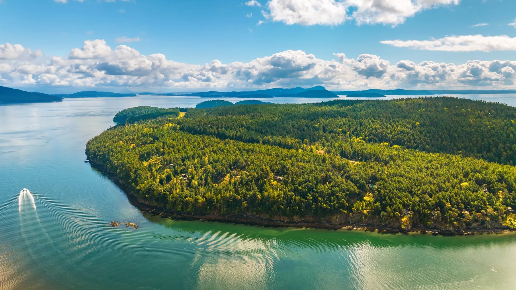Aerial view of a lush green island with dense forest surrounded by calm blue water, with a boat leaving ripples in the water.