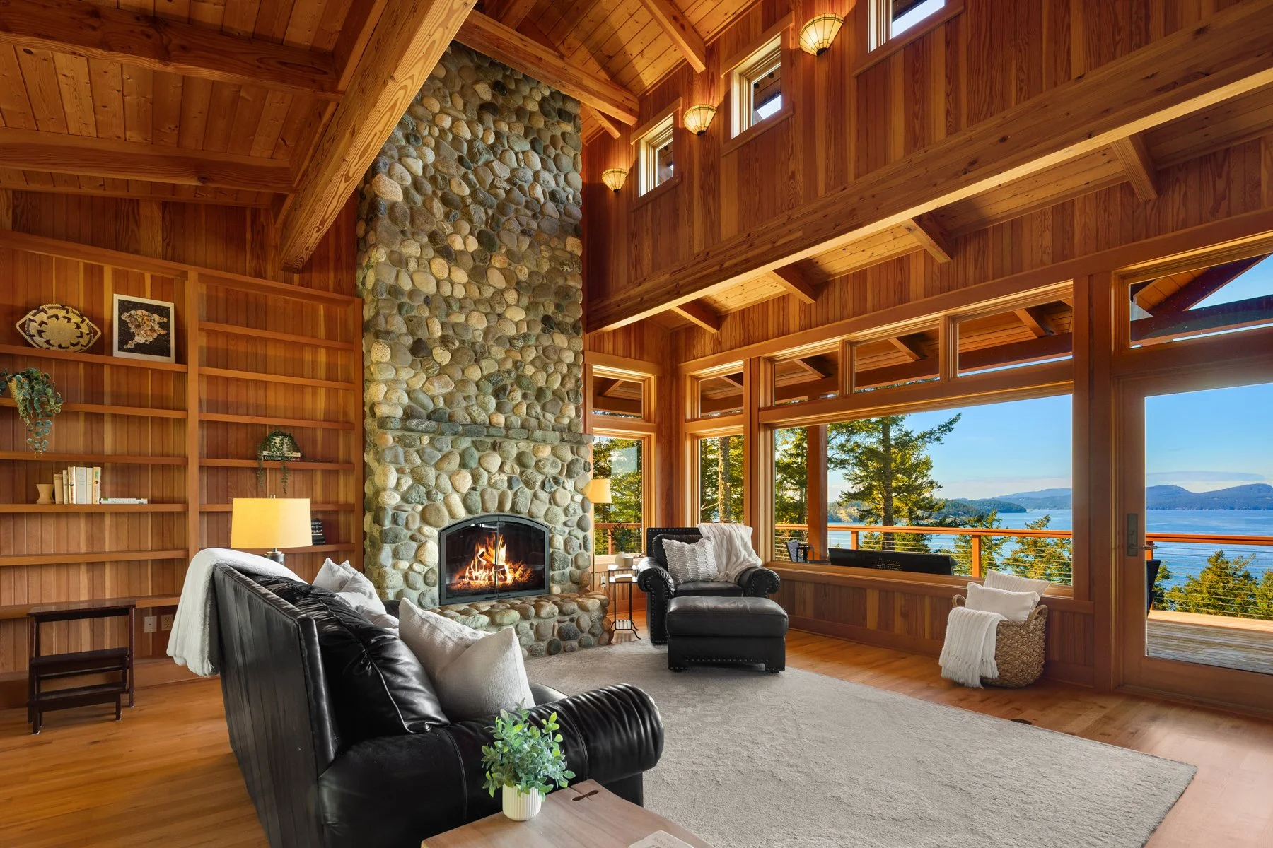 A spacious living room with wooden walls and ceiling, large windows offering a lake view, a stone fireplace with a fire, black leather sofas with pillows, a wooden bookshelf, and decorative items.
