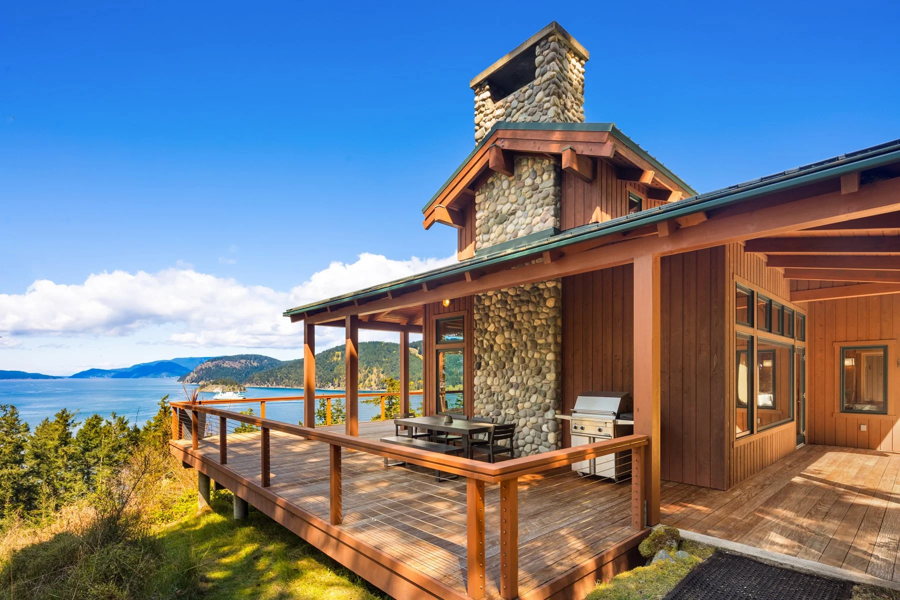 A house with a wooden deck overlooking a body of water and forested hills, featuring a stone chimney and windows.
