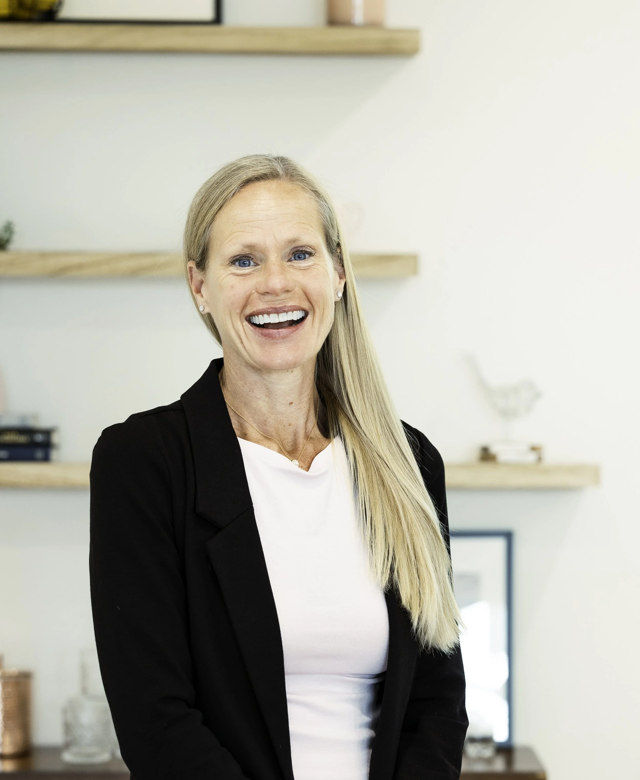 A smiling woman with long blond hair, wearing a black blazer over a white top, standing indoors with bookshelves and framed pictures in the background.