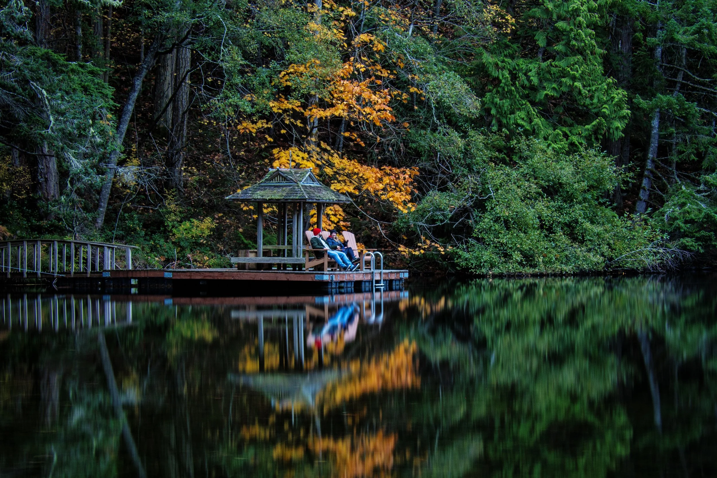 A pond in a forest with a wooden gazebo and two people sitting on a bench inside, surrounded by trees with autumn leaves.