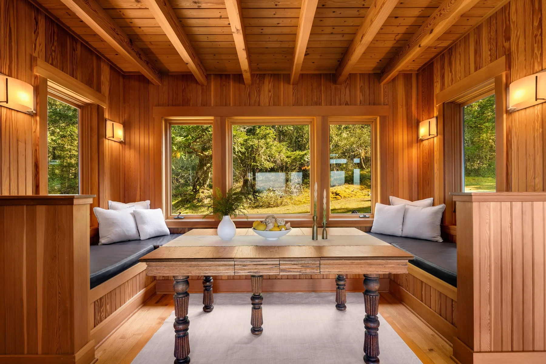 A cozy wooden dining nook with built-in bench seating on both sides, cushions, a rustic wooden table, a white vase with greenery, and a bowl of lemons. Large windows with a view of greenery and trees outside, warm wall-mounted lighting, and exposed w