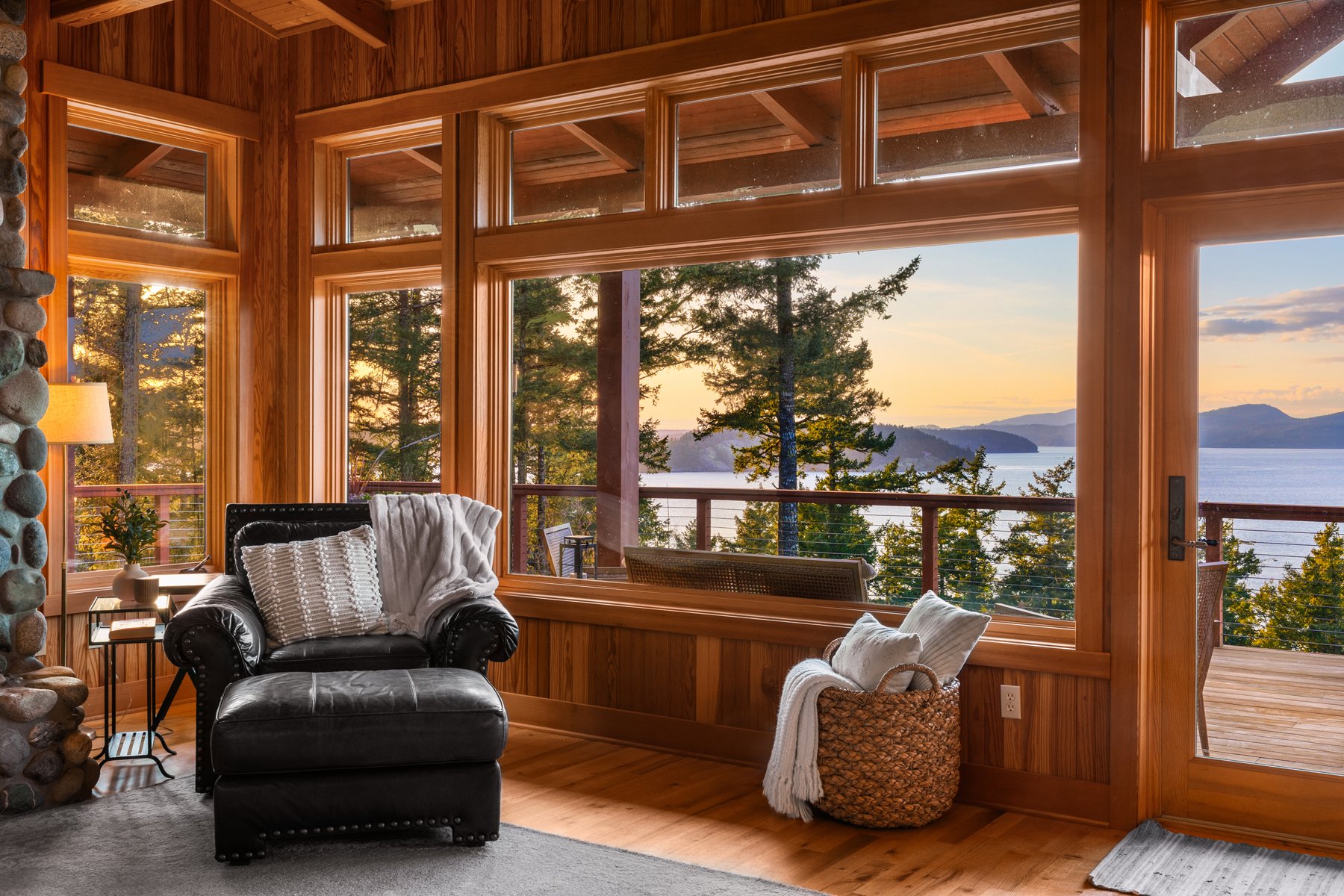 Living room with large wooden-framed windows overlooking a forest and lake at sunset. There is a black armchair with white pillows, a side table, a lamp, a woven basket with a blanket, and a balcony visible outside.