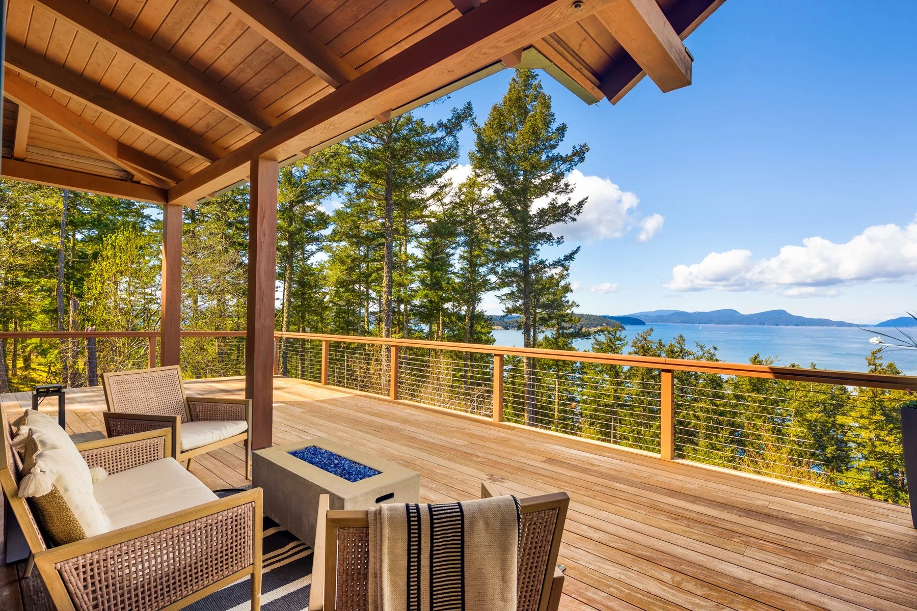 A wooden deck with outdoor furniture, overlooking a forest, a body of water, and distant mountains under a blue sky with white clouds.