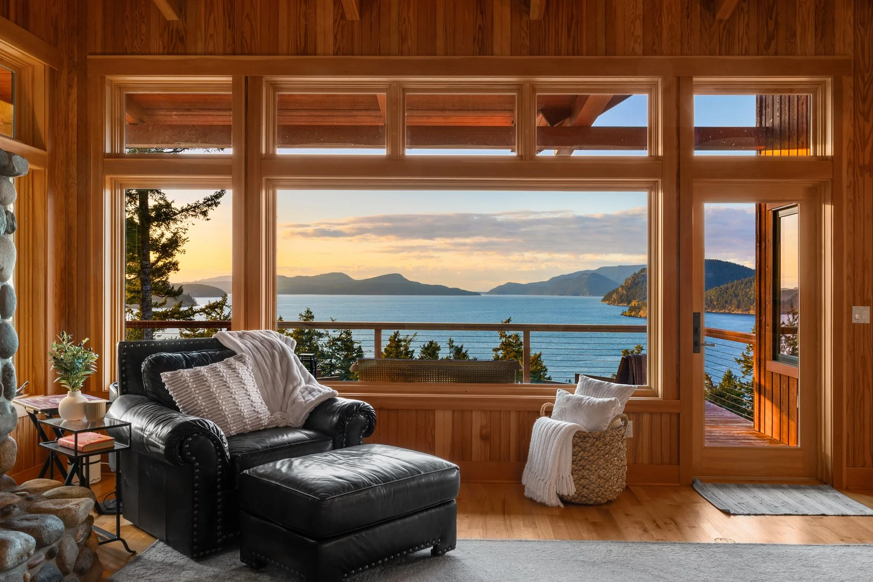 Living room with large windows showing a scenic view of a lake, mountains, and trees at sunset. The room has a black leather sofa with white pillows and a throw blanket, a side table with a plant and books, and a wicker basket with a blanket near the door.