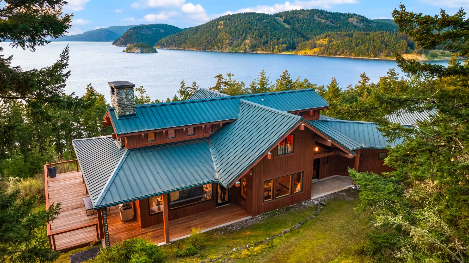 A wooden house with a green metal roof situated among trees overlooking a large lake with forested hills in the background during daytime.