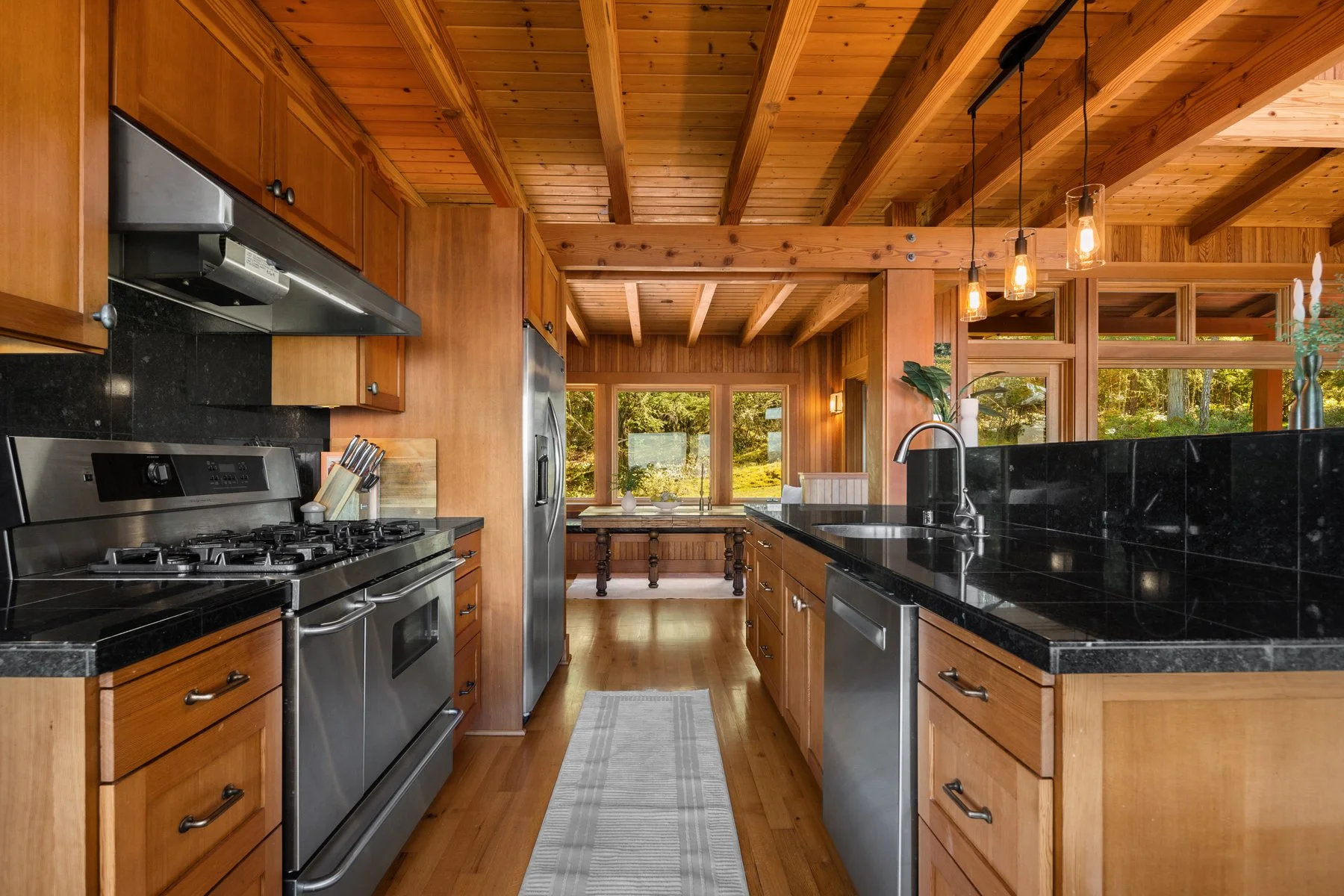 Modern kitchen with wooden cabinets and ceiling, black granite countertop, stainless steel appliances, and large windows showing a lush outdoor scene.