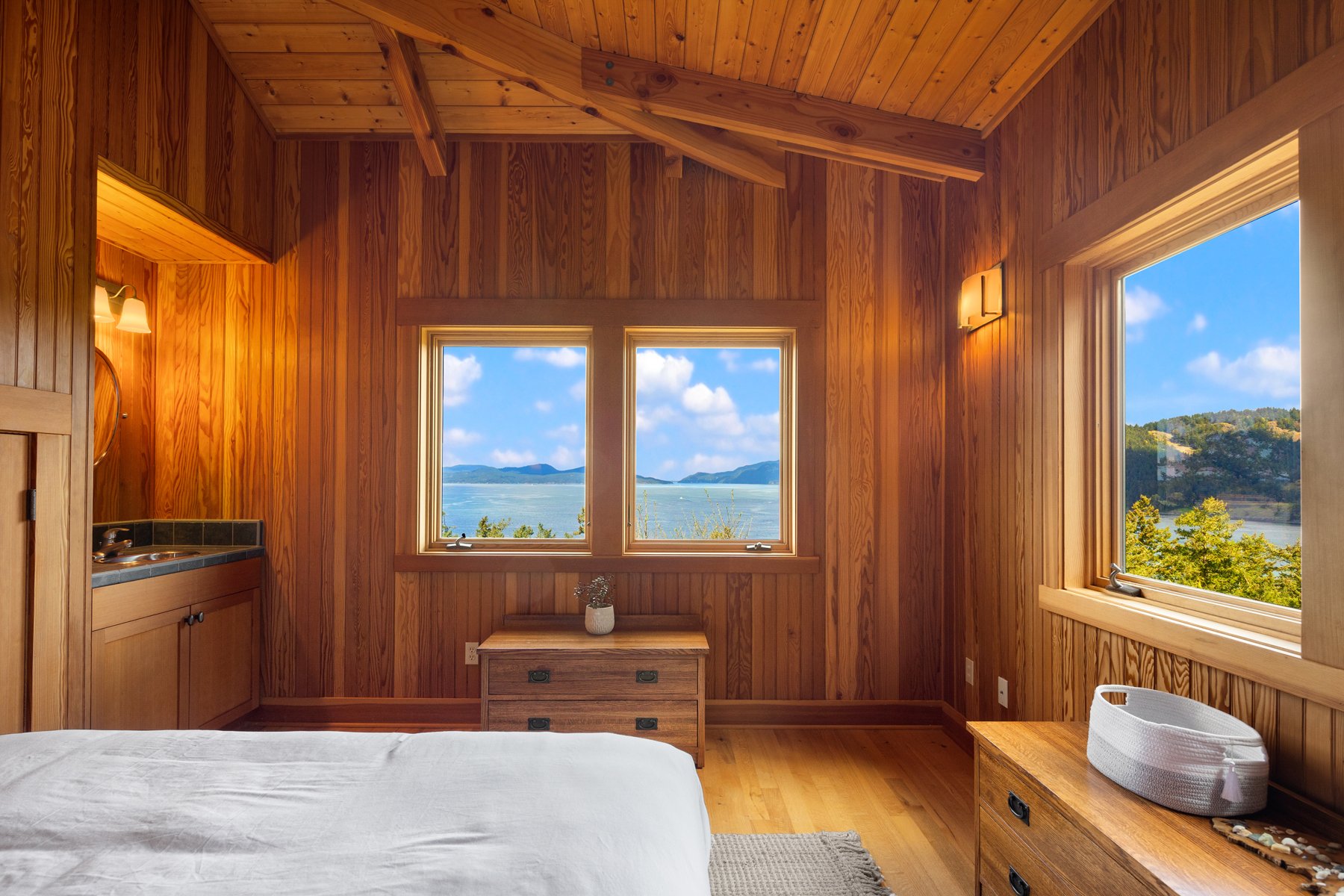 Interior of a wood-paneled bedroom with two windows showing a lake and mountains, wooden furniture, a bed, a small dresser with a plant, and scenic views outside.