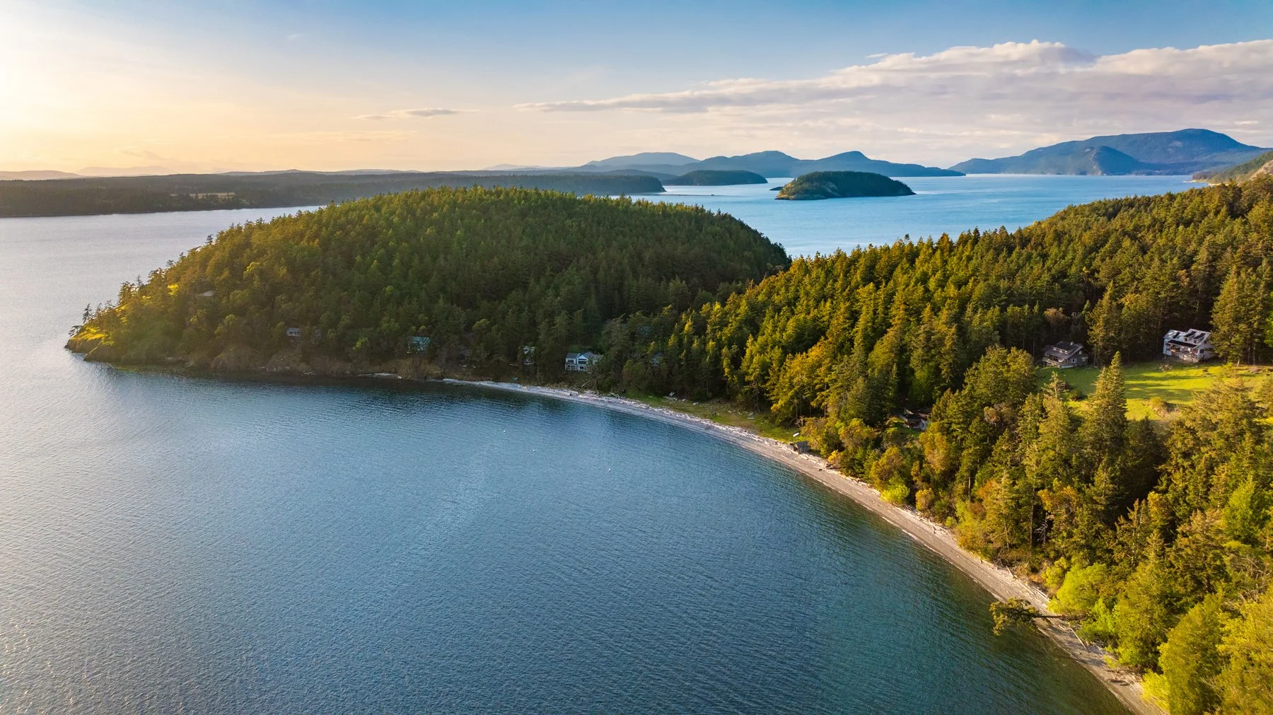 Aerial view of a large body of water with forested islands and shoreline, homes nestled among trees, distant mountains, clear blue sky, and a setting sun.