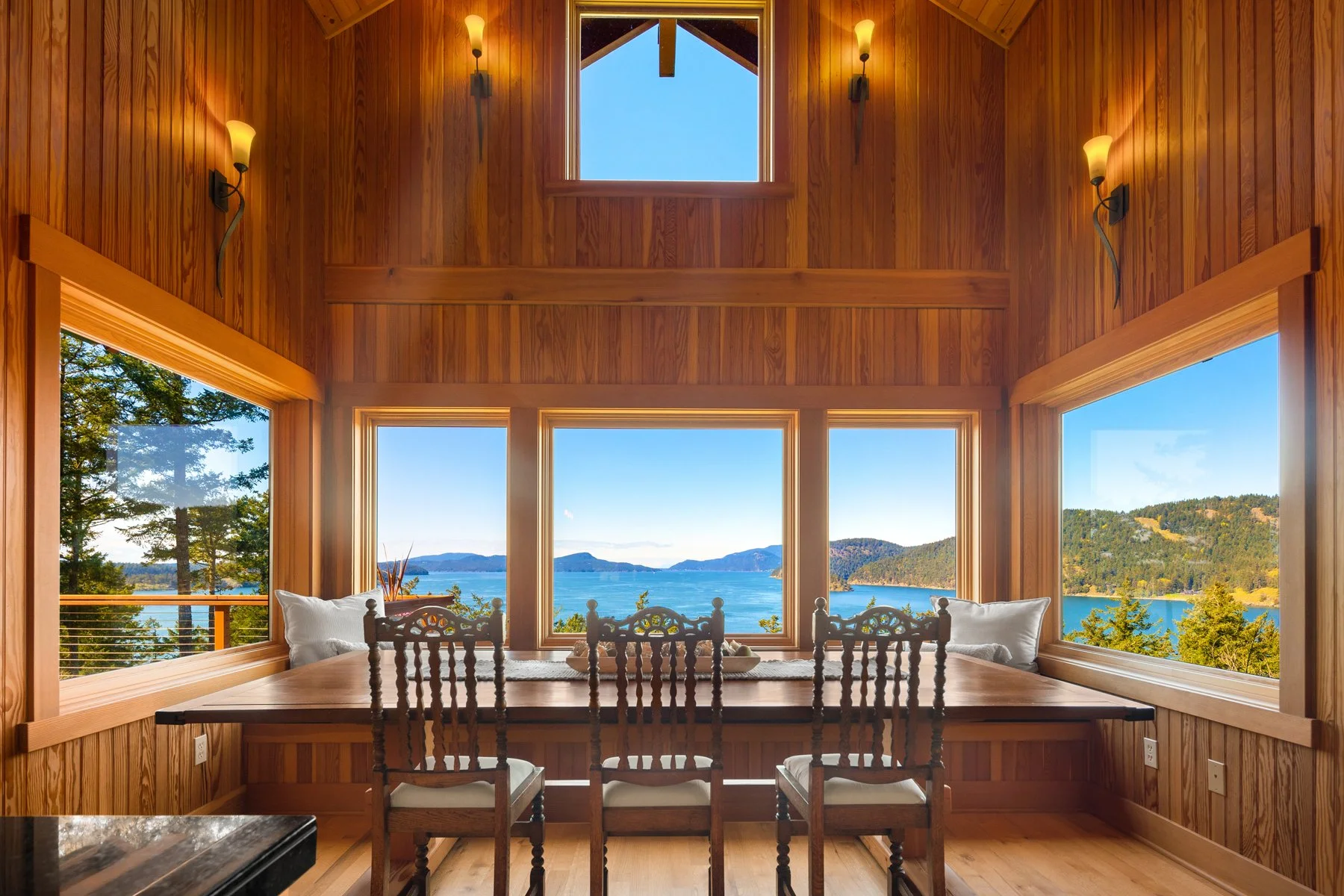 Wooden interior dining room with large windows showcasing a scenic lake and mountain view, four wooden chairs around a wooden table, pillows on the window seats, and wall sconces lighting.