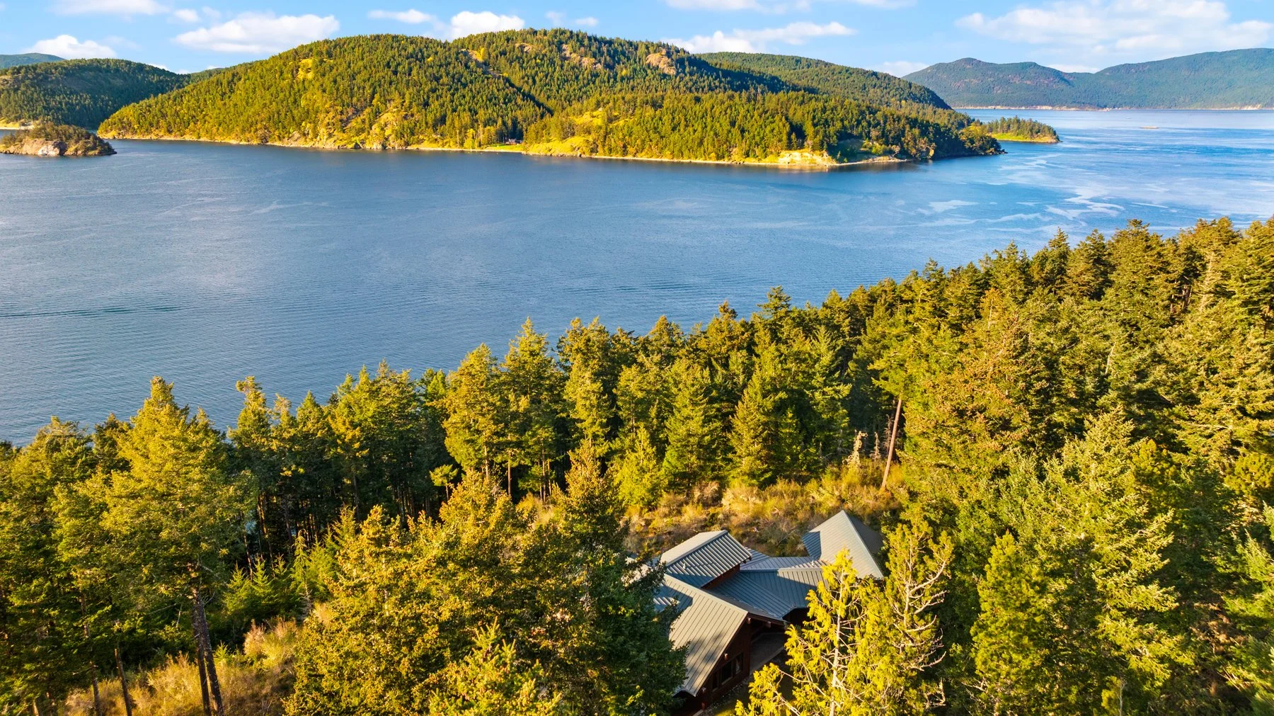 A scenic aerial view of a lake surrounded by dense green forests and rolling hills, with rooftops of houses visible among the trees.