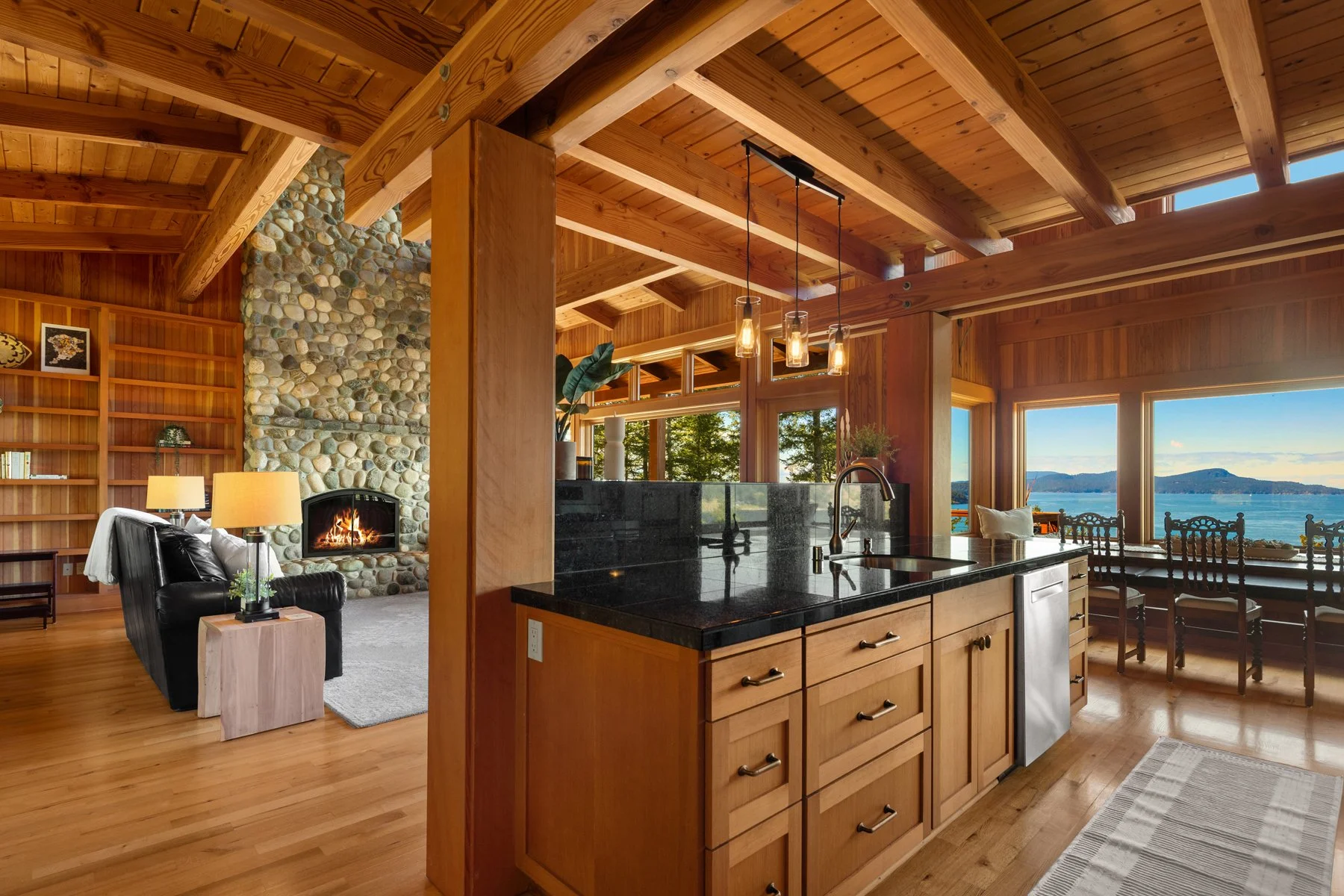 Open-concept living space with a stone fireplace, wooden ceiling and beams, black leather armchair, side table with lamp, kitchen island with black countertop, and large windows showcasing a lake view with mountains.