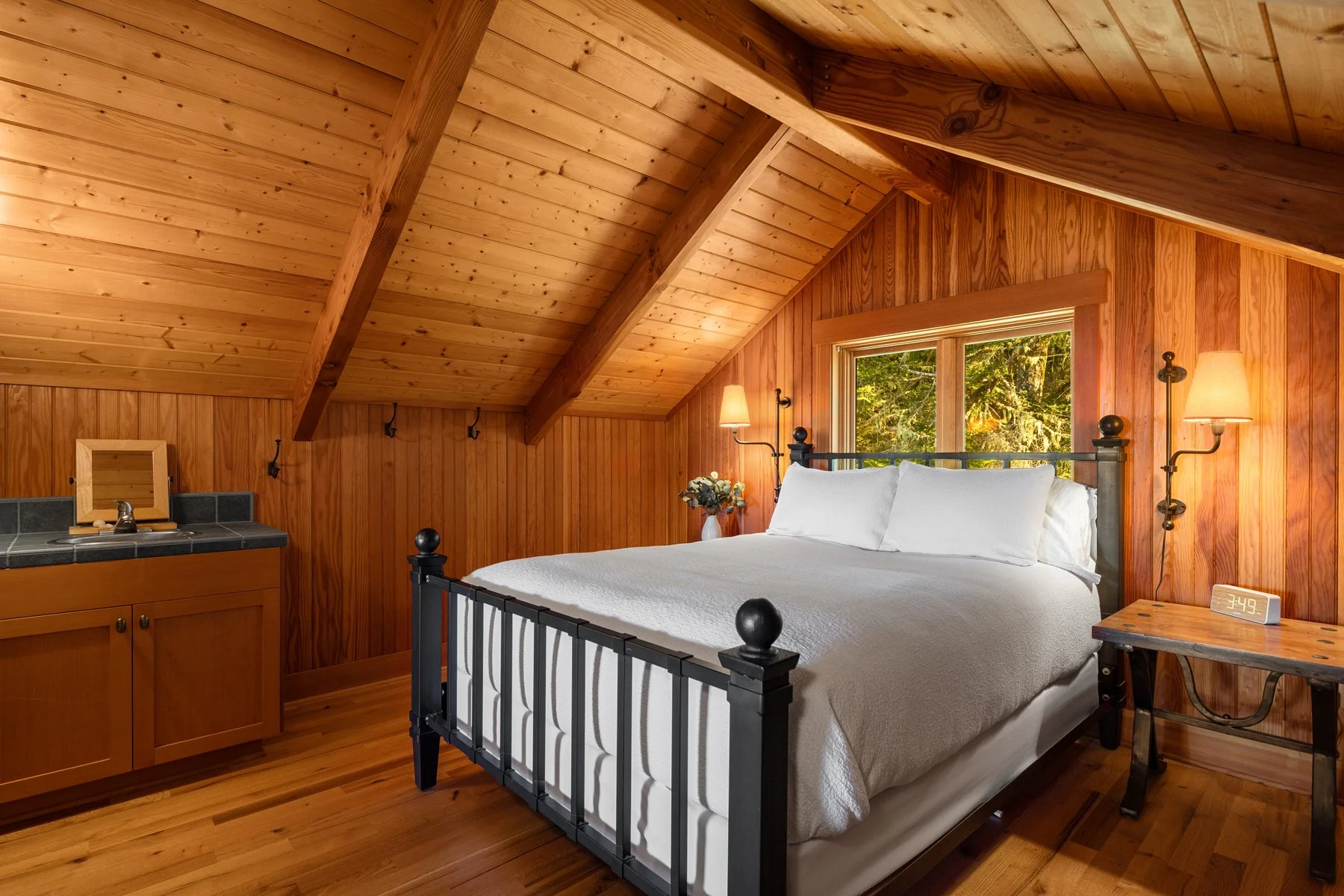 A cozy bedroom with wood-paneled walls and sloped ceiling, featuring a black metal bed with white bedding, two wall-mounted lamps, a small bedside table with a digital clock, and a window with greenery outside.