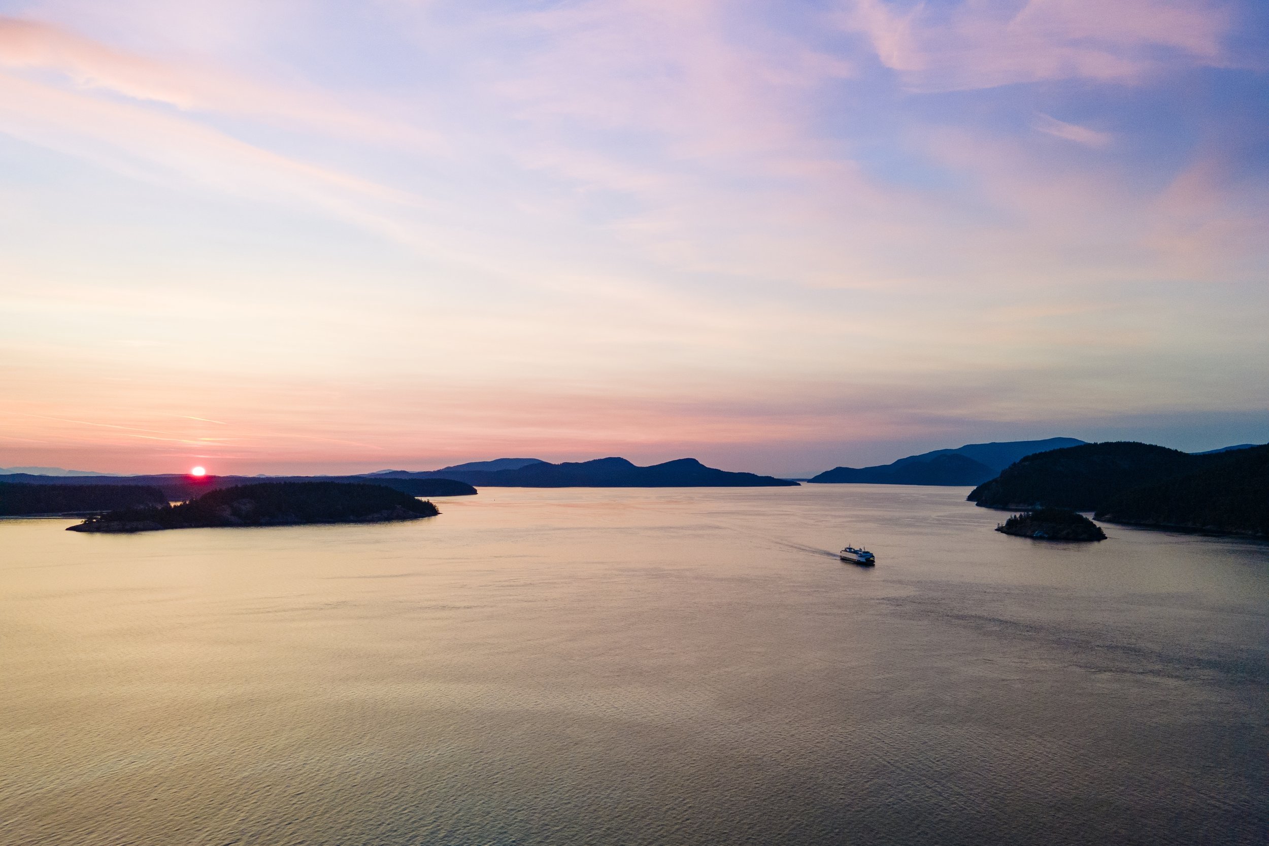 A scenic view of a large body of water at sunset with hills in the background and a boat sailing.