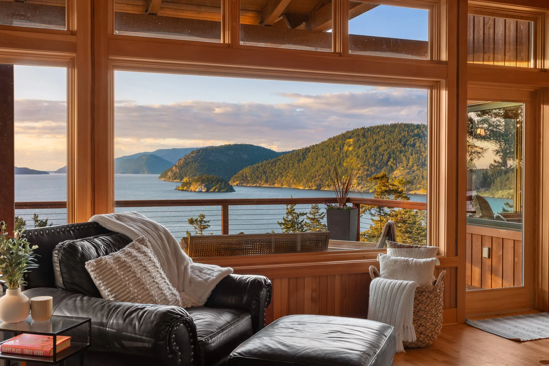 Living room with large windows showing a scenic view of a lake, mountains, and trees; black leather sofa with a white blanket, a side table with books and vases, and indoor plants.