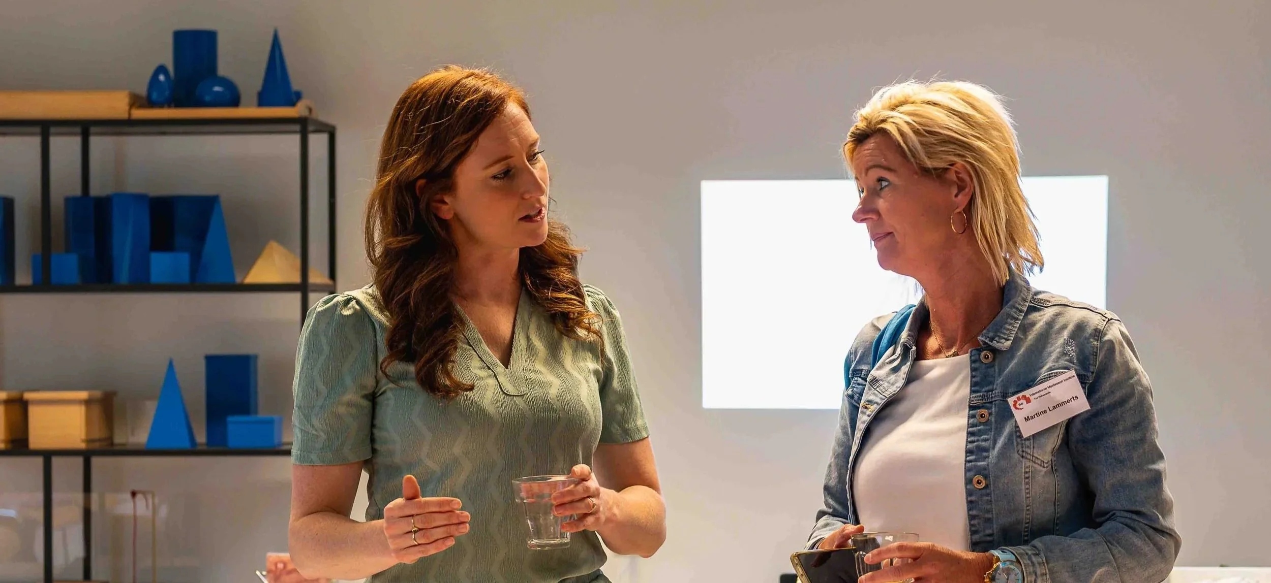 Two women talking at a social gathering, holding drinks, standing indoors near a shelf with blue decor items and a bright window in the background.