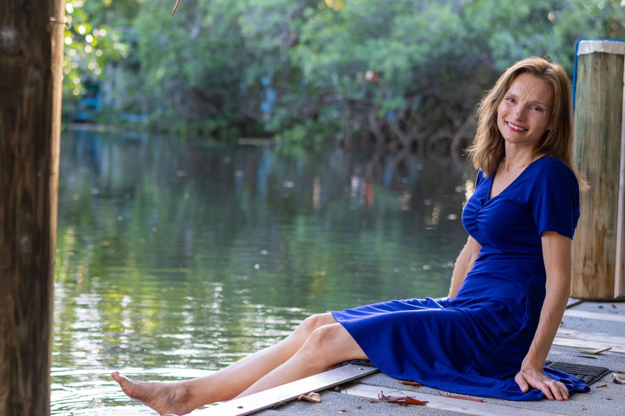 A woman sitting by a river, stretching her legs, wearing a blue dress with a background of lush green trees and water.