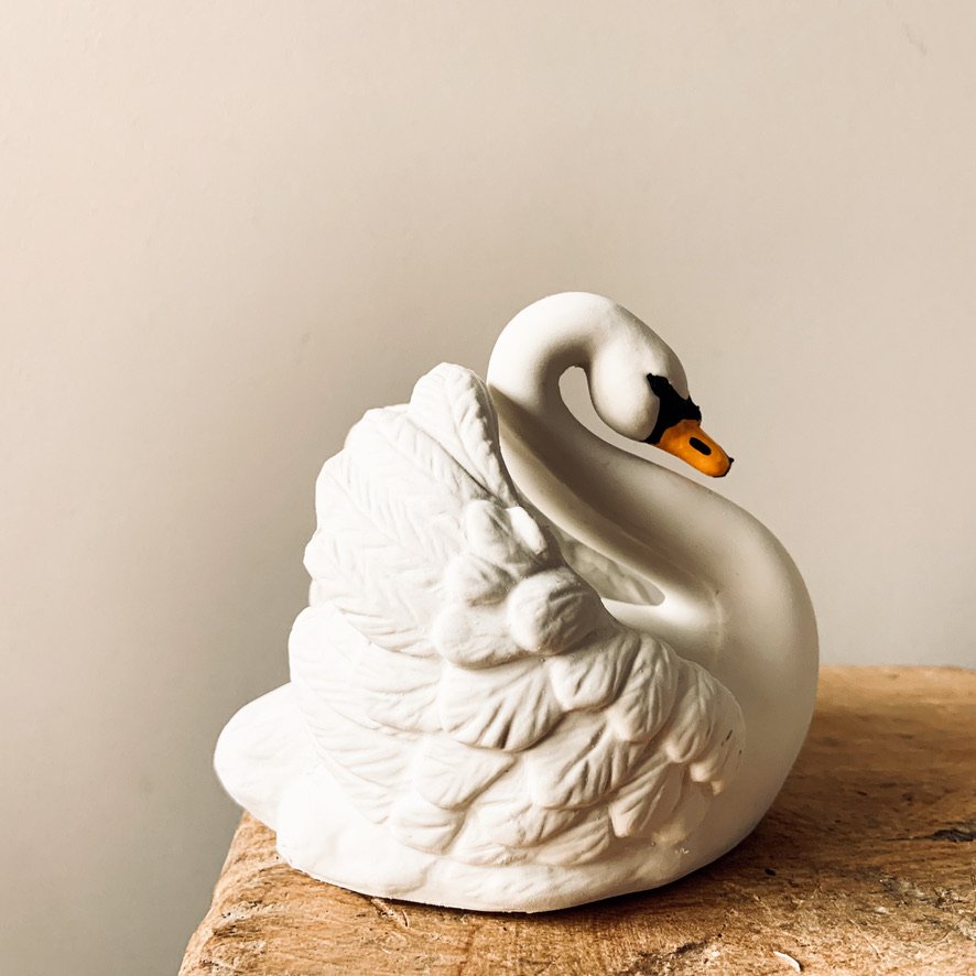 A decorative ceramic swan figurine with a textured wing and a smooth neck, sitting on a wooden surface, with a plain light-colored wall background.