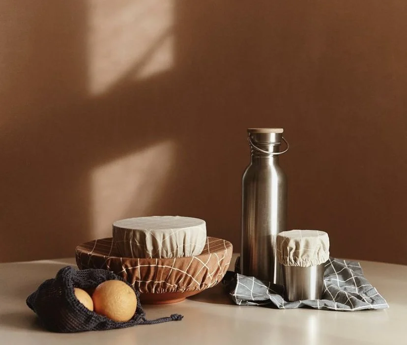 A still life composition with cheese covered in cloth, oranges in a black net bag, and stainless steel bottles on a table with a checkered cloth, against a warm brown background with shadow patterns.
